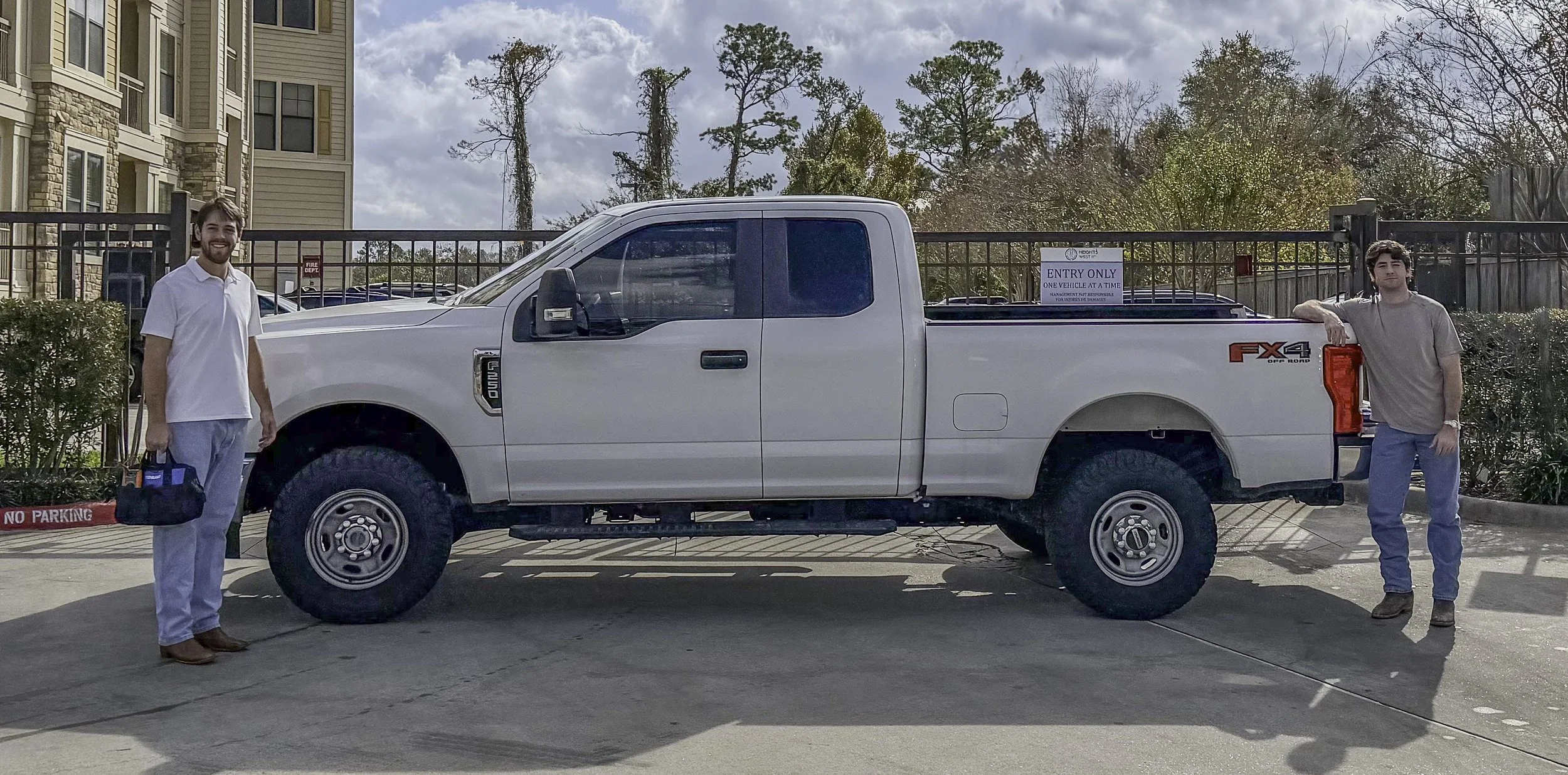 Two young men standing on either side of a white Ford FX4 pickup truck, parked in a lot with a sign indicating only one vehicle at a time entry. The man on the left is dressed in a white shirt and light-colored pants, holding a bag, and smiling. The man on the right is dressed in a gray shirt and darker pants, with one arm resting on the truck's tailgate. In the background, there are trees, apartment buildings, and a partly cloudy sky.