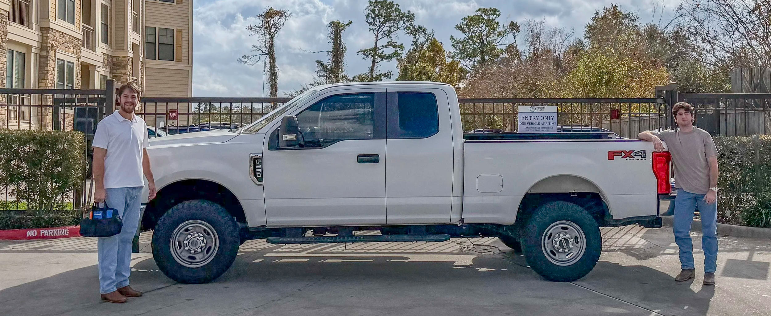 Two young men standing next to a white pickup truck with large tires in a parking lot with apartment buildings and trees in the background.