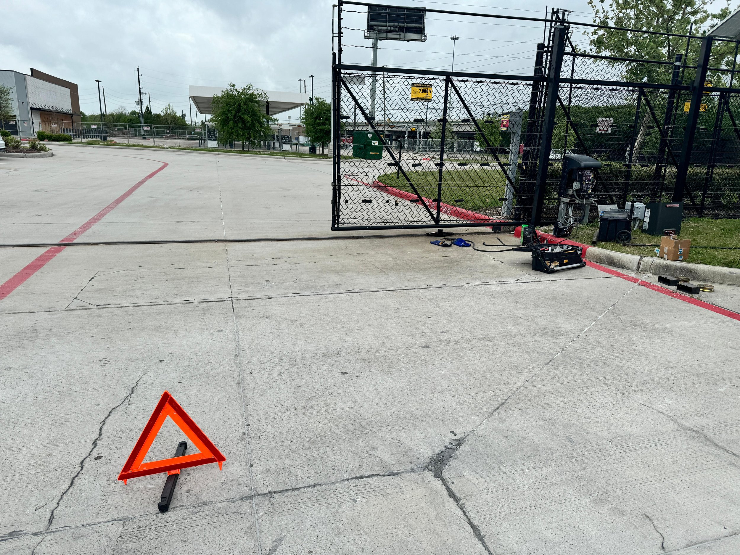 Parking lot with a temporary traffic sign and tools, black fence, and some equipment on the ground, under an overcast sky.