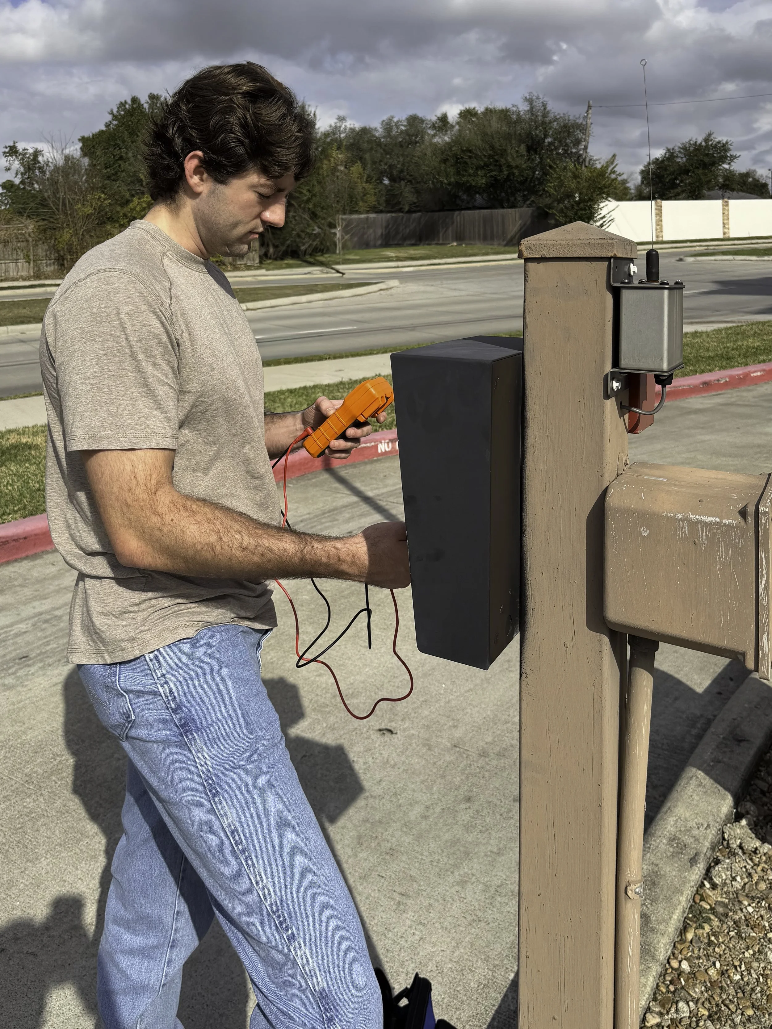 A man standing outside on a concrete sidewalk using a digital multimeter to test electrical wiring on a wooden post near a parking lot.