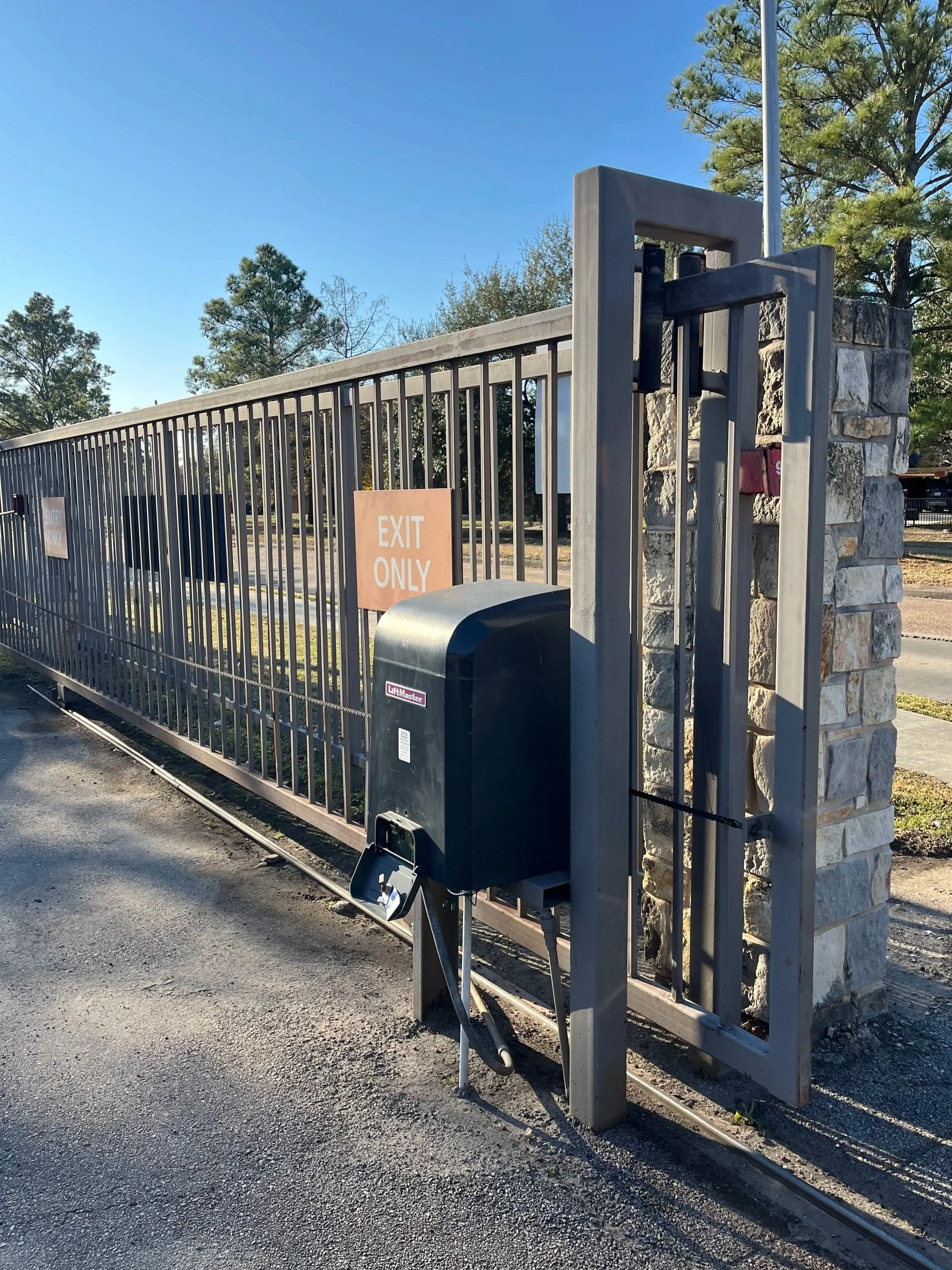 A metal gate with a stone pillar, signs that say 'Exit Only', and a trash bin. There are trees and a clear blue sky in the background.