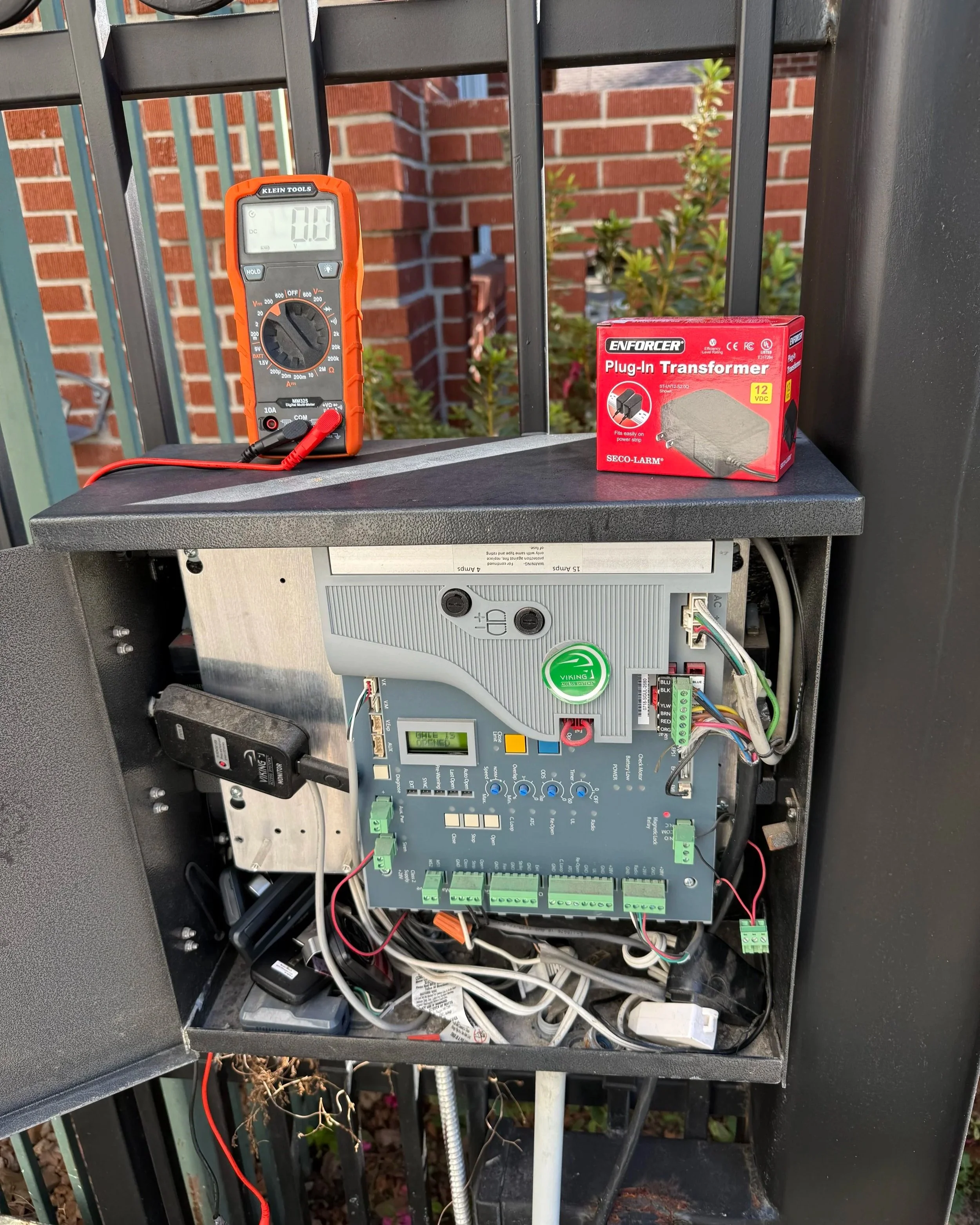 Open electrical box with multimeter, wires, circuit board, and a small red box labeled 'Enforcer Plug-In Transformer' outside on a metal railing with brick wall and plants in the background.