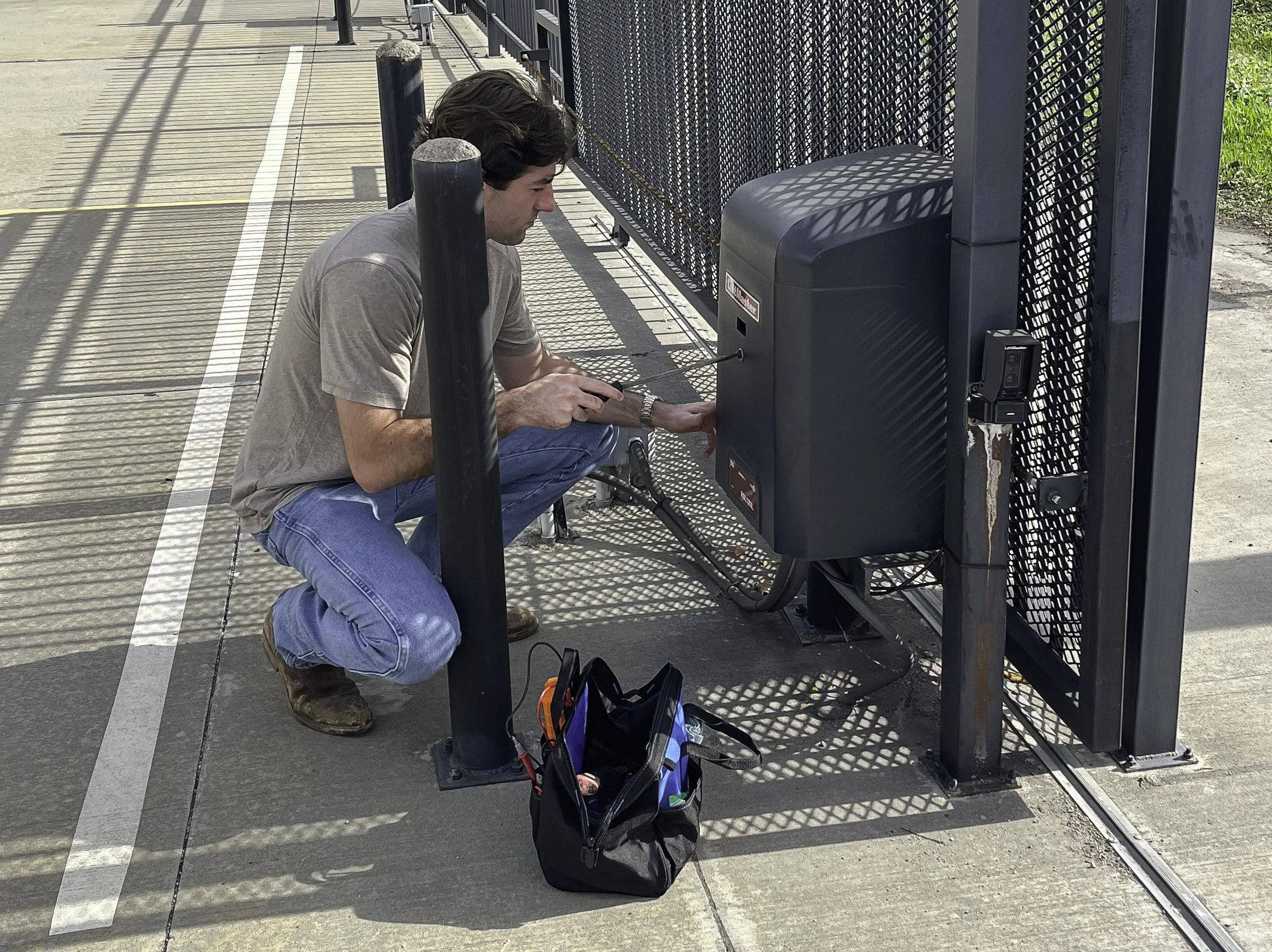A man crouching next to a bike lock station, using a phone or device, with a black bag on the ground beside him. The scene is outdoors on a sidewalk next to a fence.