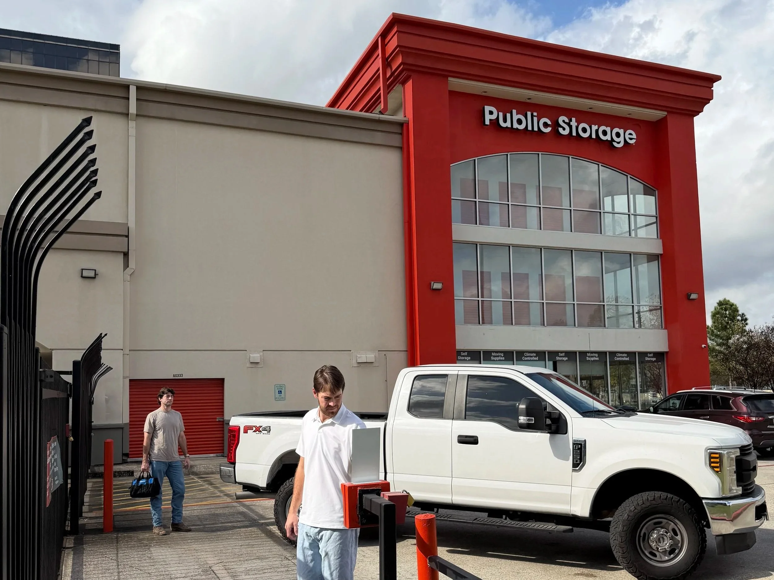 People in parking lot outside a multi-story storage facility. One person is walking away, another is near a white pickup truck, and a third person is looking down, possibly at a phone or device.