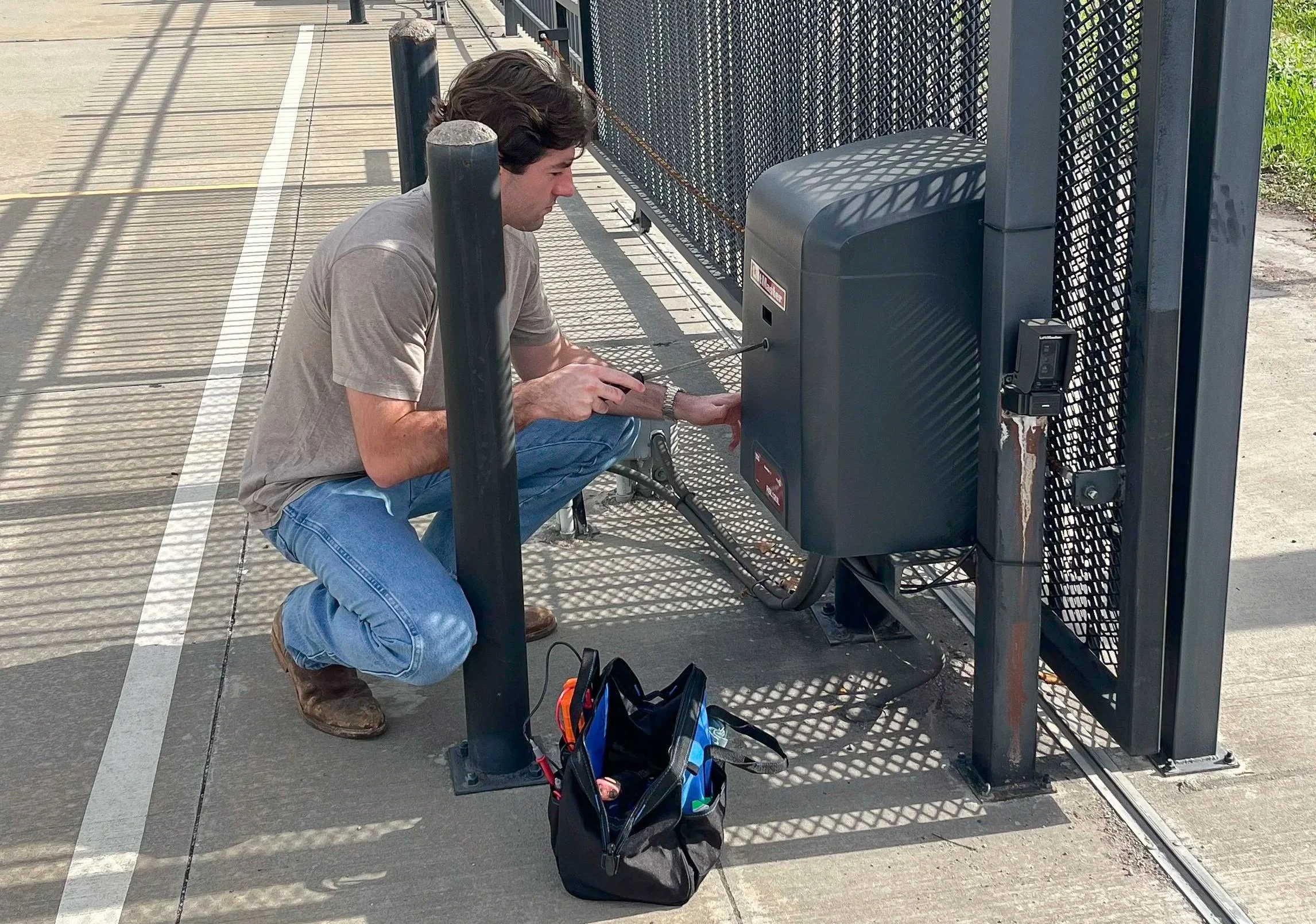 A man crouched down outside at a fence, working on an electrical box or control panel, with tools and a black bag nearby.