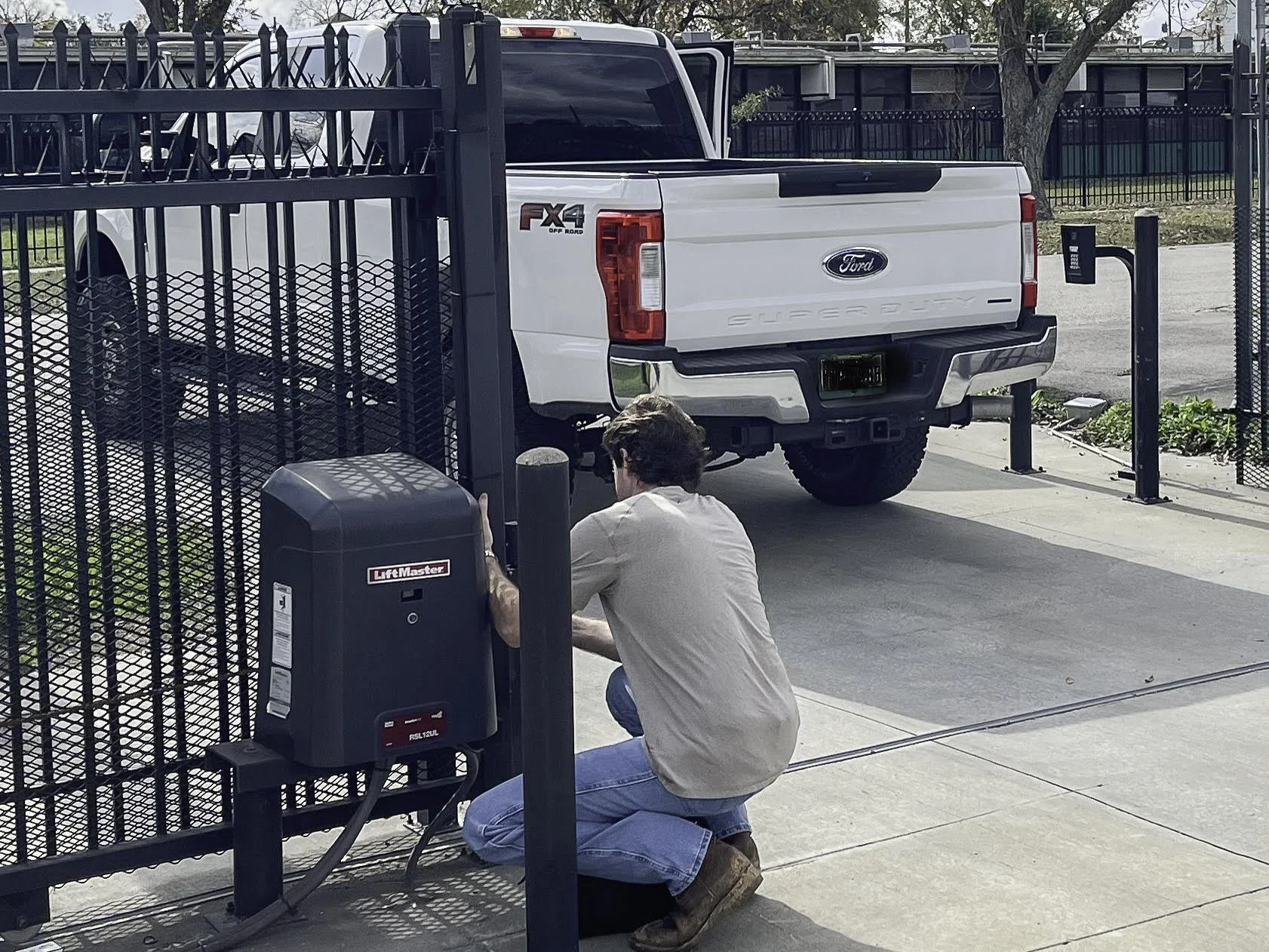 A man kneeling outside a gate, working on a LiftMaster automatic gate opener system. A white Ford Super Duty pickup truck is parked nearby.