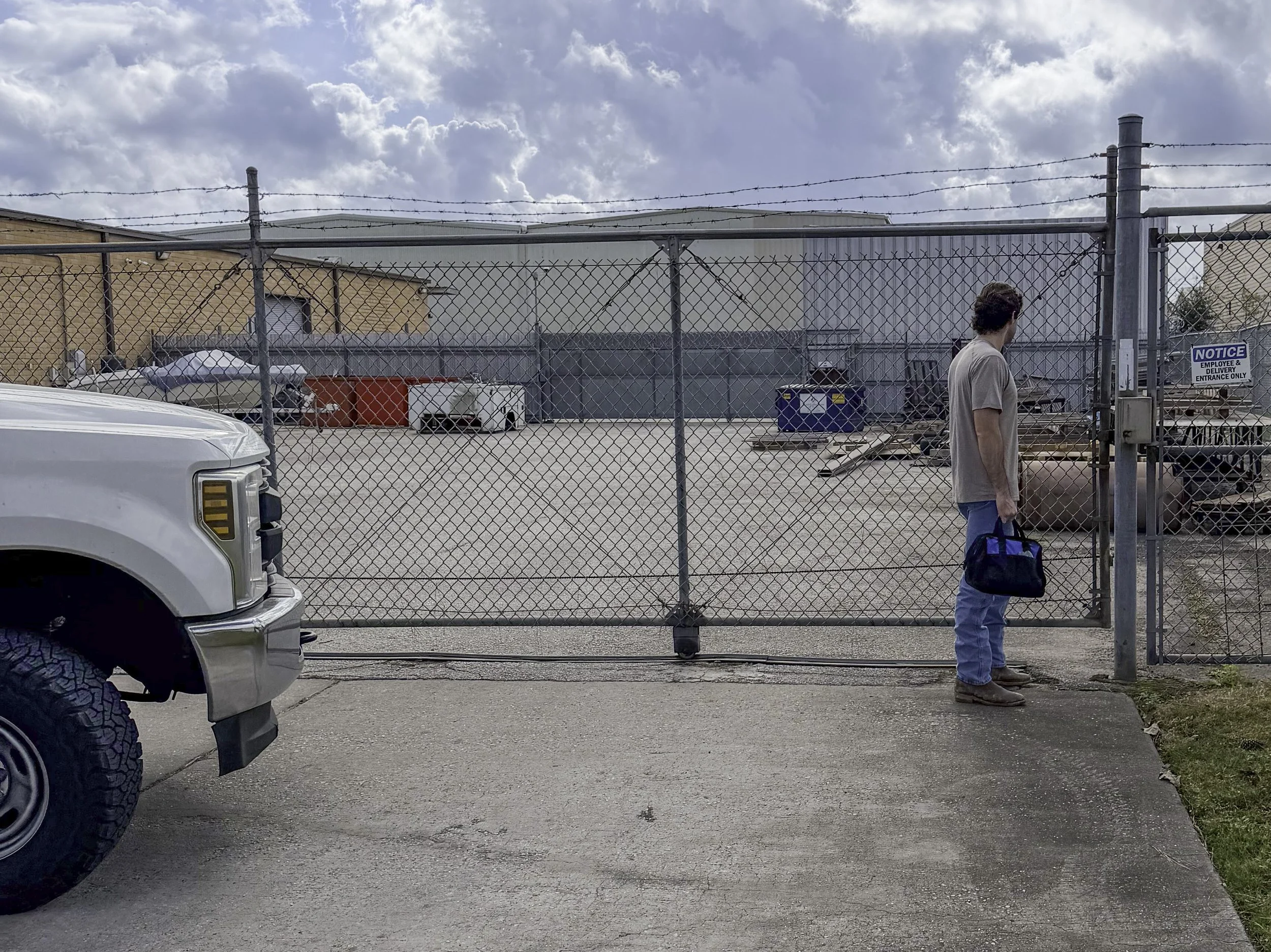 A man standing outside a fenced lot, holding a blue bag, with a white truck in the foreground and a notice sign on the fence.