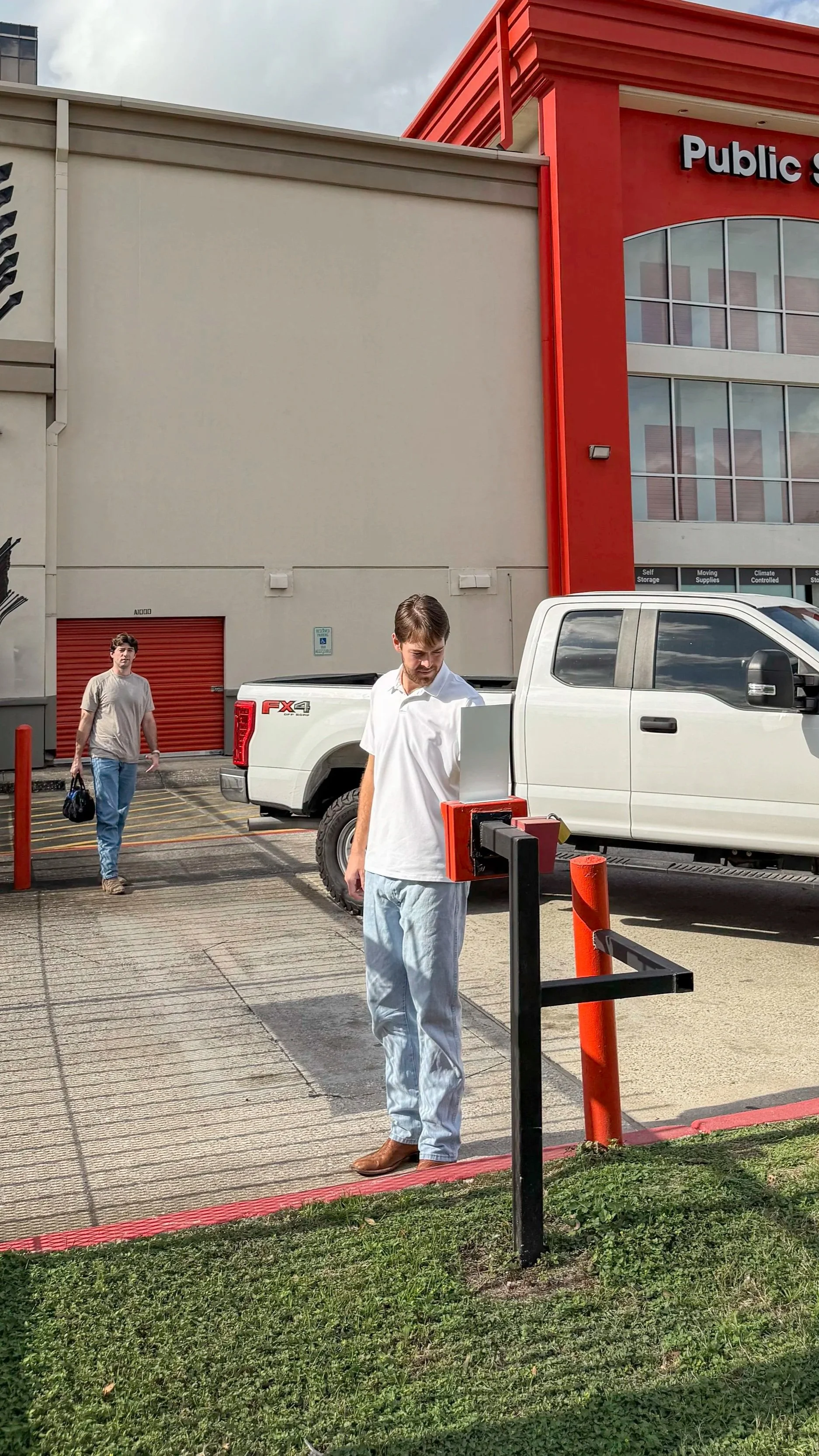 Two young men outside a retail store, one using a pay station, the other carrying a bag. One person is in a white shirt and light pants, the other in a gray shirt and jeans. The store has a red facade with large windows and a sign that reads 'Public.' There are trucks parked nearby and parking barriers in front of the store.
