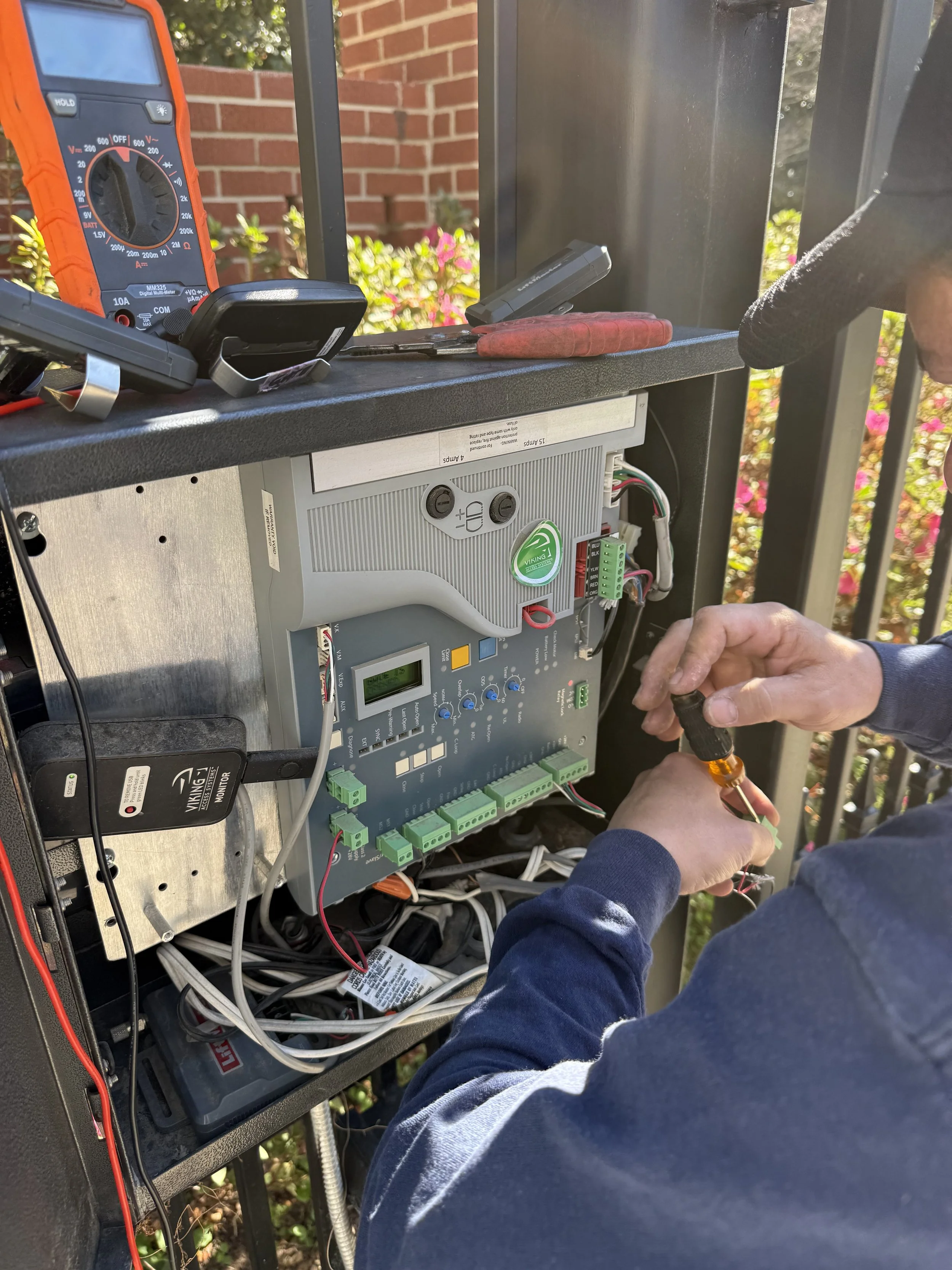 Technician working on electrical equipment outdoors, using tools and a multimeter, with various wires and components visible inside an open control panel.