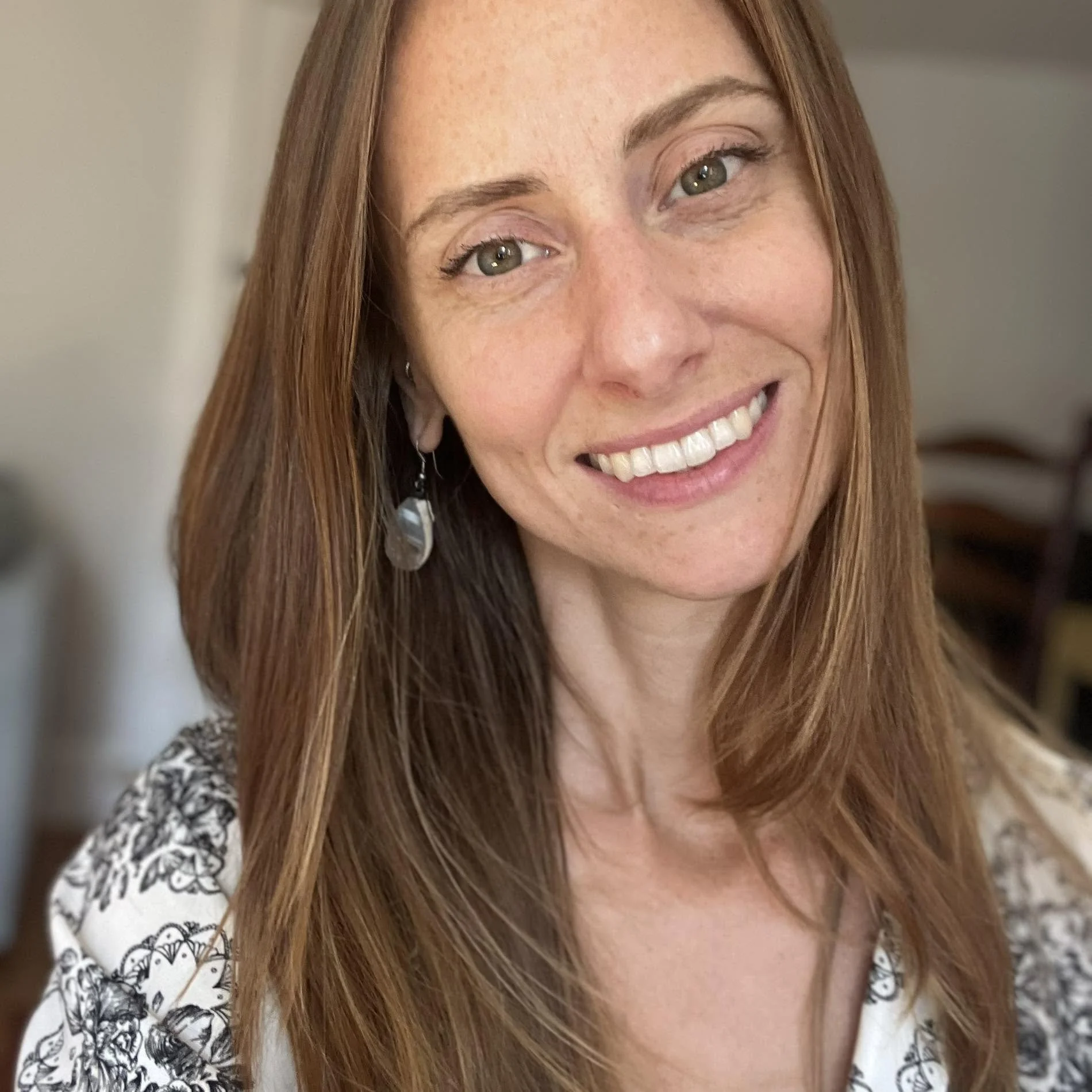 A woman with long, light brown hair smiling at the camera, wearing a green dress with textured dots and a gold necklace with black and gold pendant.