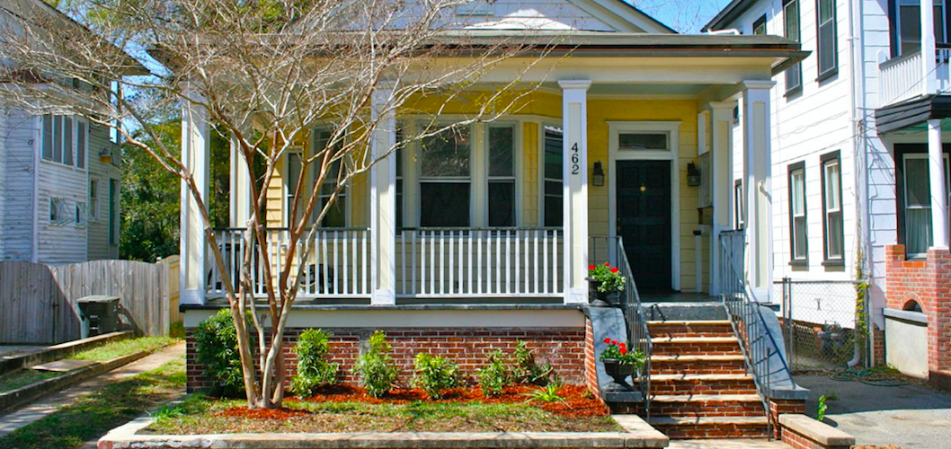 Front view of a yellow house with a front porch, brick stairs, and a small garden with plants and a tree, located in a residential neighborhood.