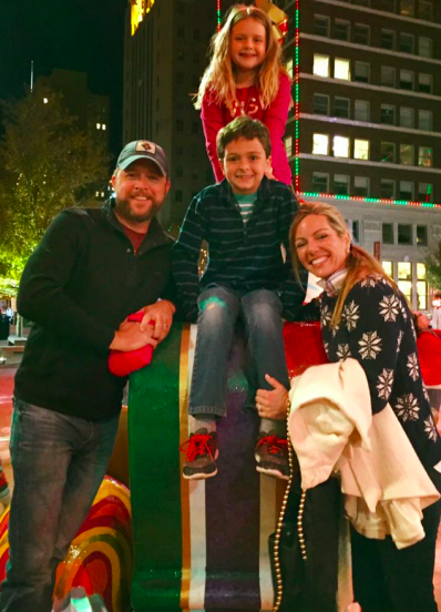 A family of four enjoying a festive night outdoors, with two adults and two children smiling while posing on a colorful holiday display near a tall building decorated with Christmas lights.