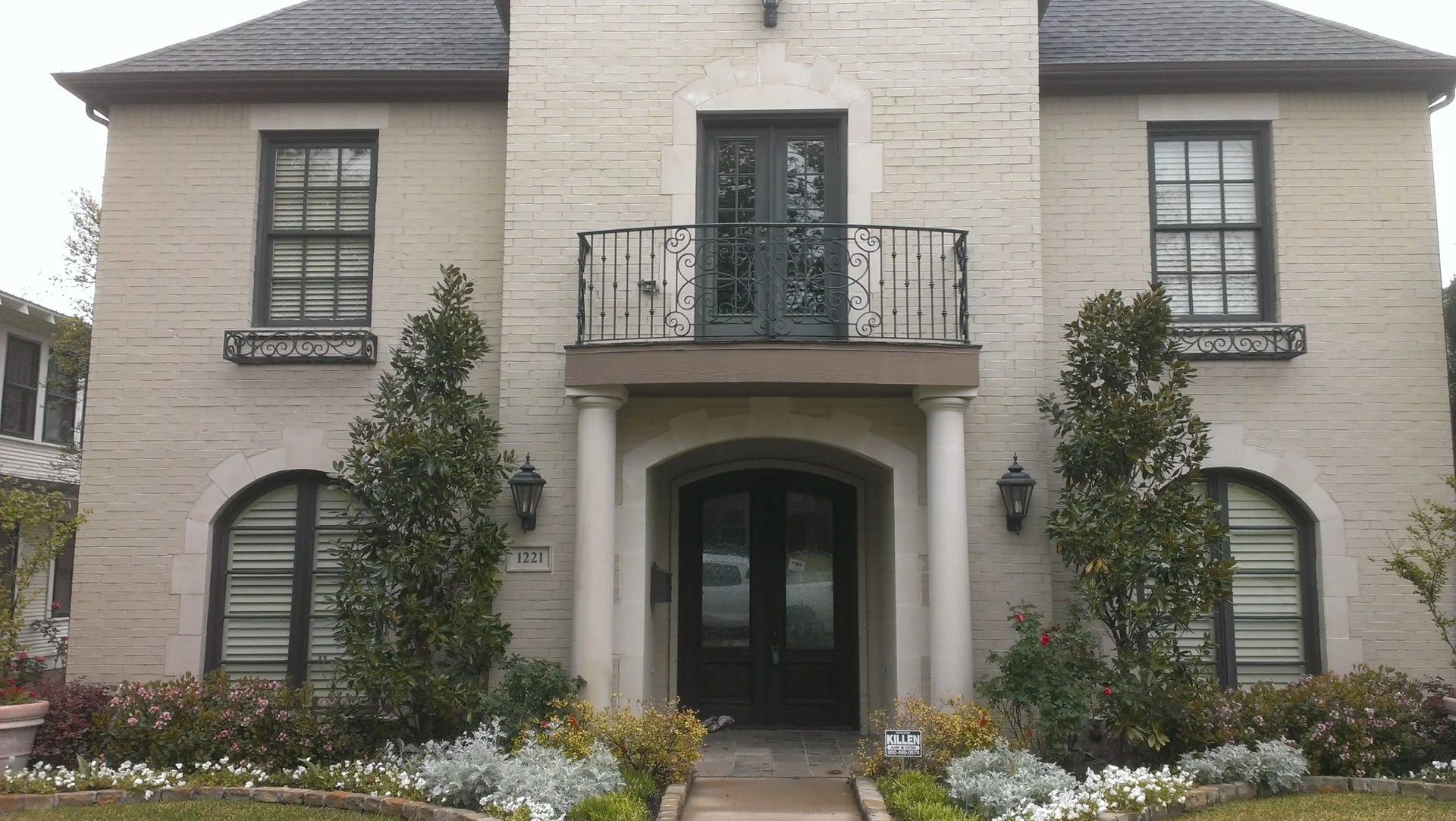 Front view of a two-story beige brick house with a black double door, white columns, and black wrought iron balcony, surrounded by landscaped gardens.