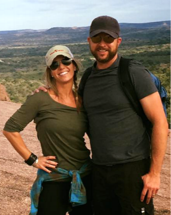 A smiling woman and man outdoors in a desert landscape, wearing hats and sunglasses, posing with their arms around each other.