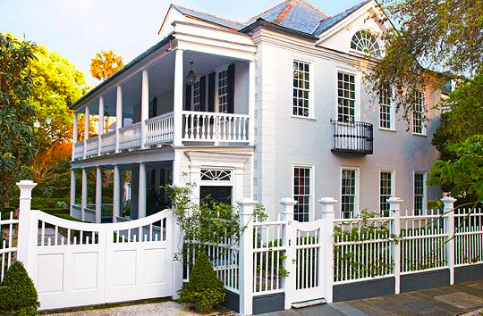 A large white house with a wrap-around porch and balcony, surrounded by a white fence and greenery.
