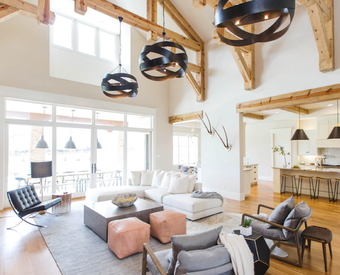 Modern living room with white sectional sofa, gray armchair, black lounge chair, and pink ottomans. Large windows and high vaulted ceiling with exposed wood beams. Contemporary black pendant lights hanging from ceiling.