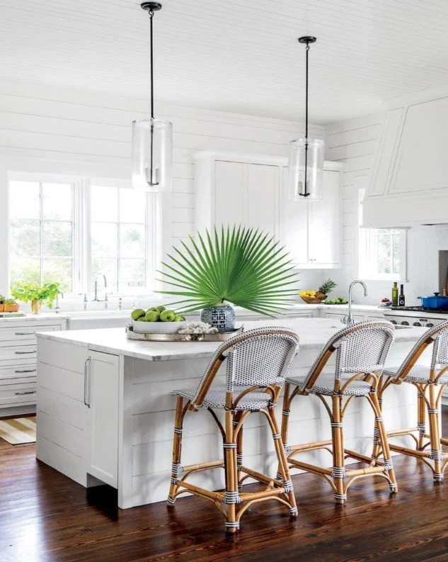 Bright white kitchen with island, pendant lighting, and wicker bar stools, decorated with a large green palm leaf centerpiece.