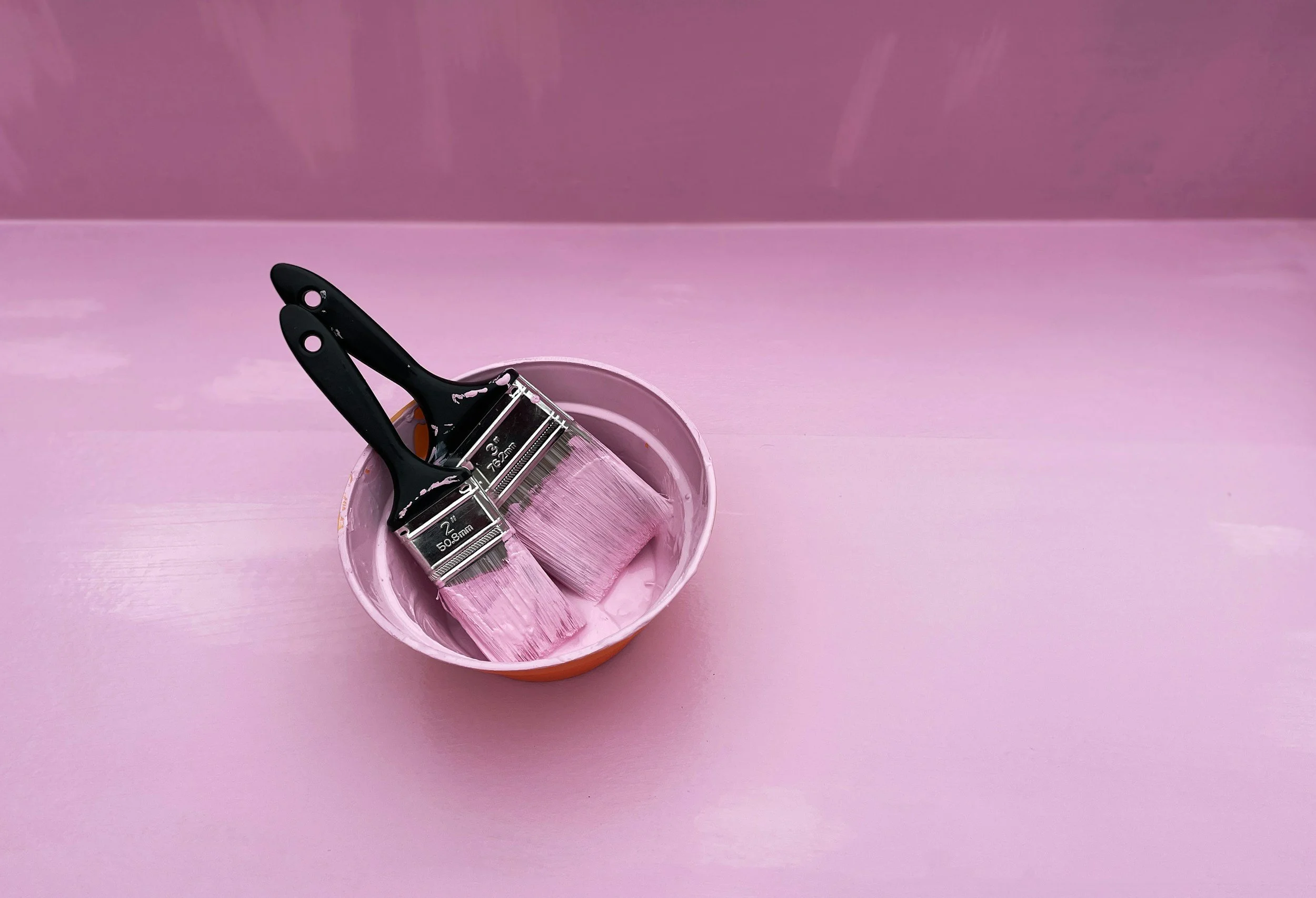 Two paintbrushes with black handles and pink bristles dipped in pink paint sitting in a small pink bucket on a pink wooden surface.
