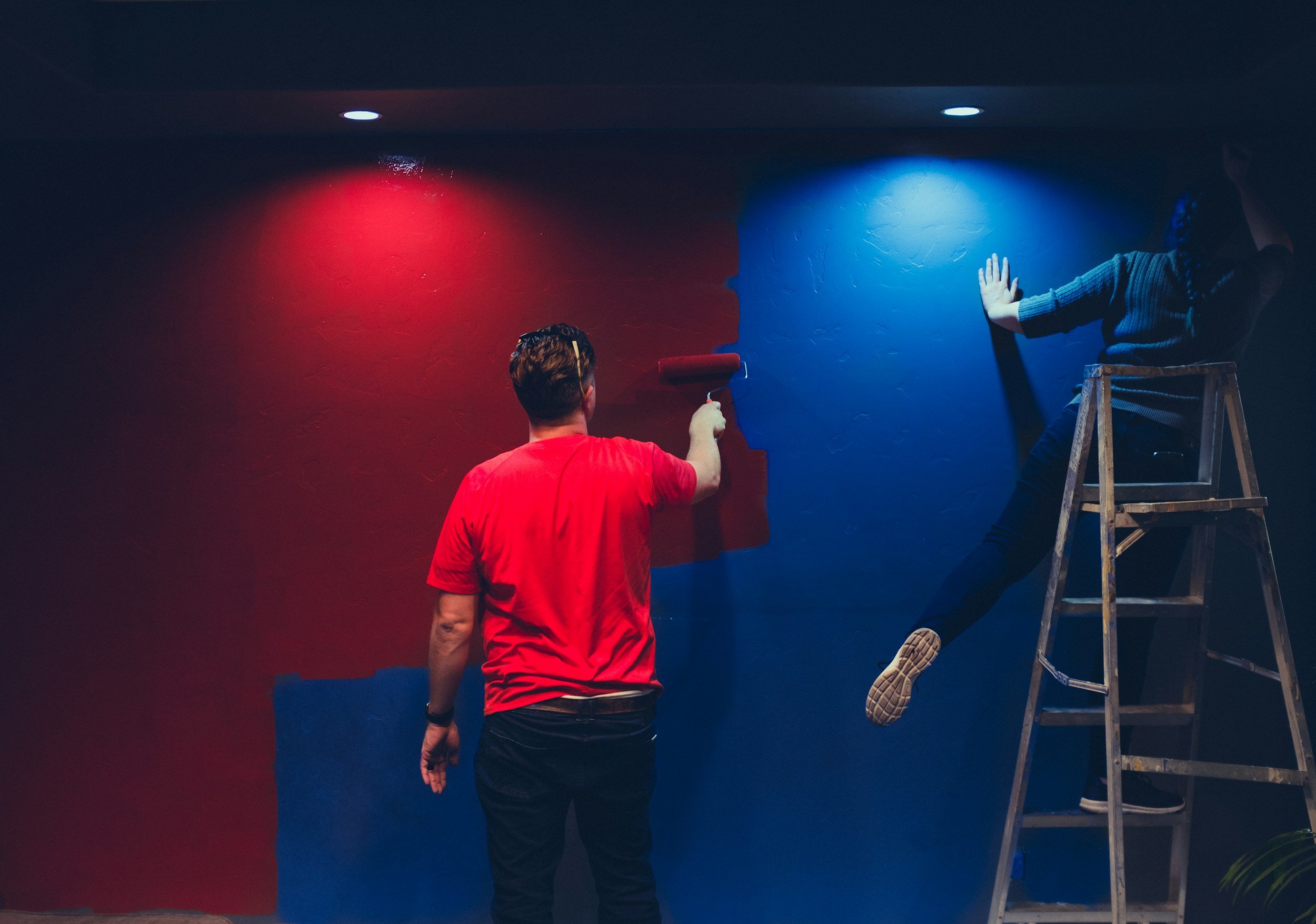 Two people painting a wall: one person in a red shirt using a roller to paint the wall red, and another person in a gray sweater on a ladder painting the wall blue.