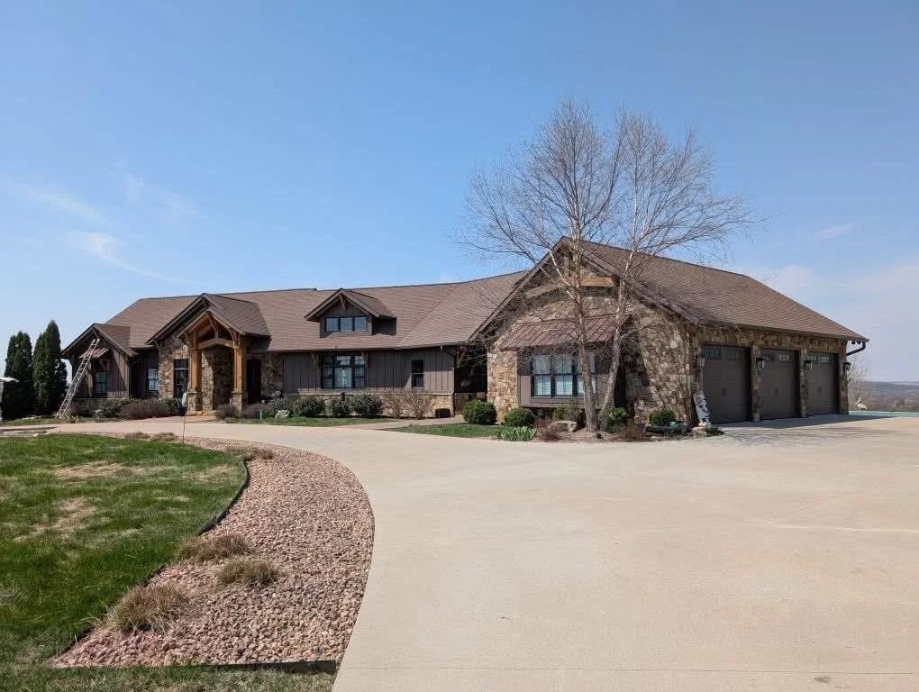 A large residential house with a brown roof, stone accents, and wooden siding. It has a three-car garage, a leafless tree in front, and a gravel-lined driveway, with a lawn and clear blue sky.