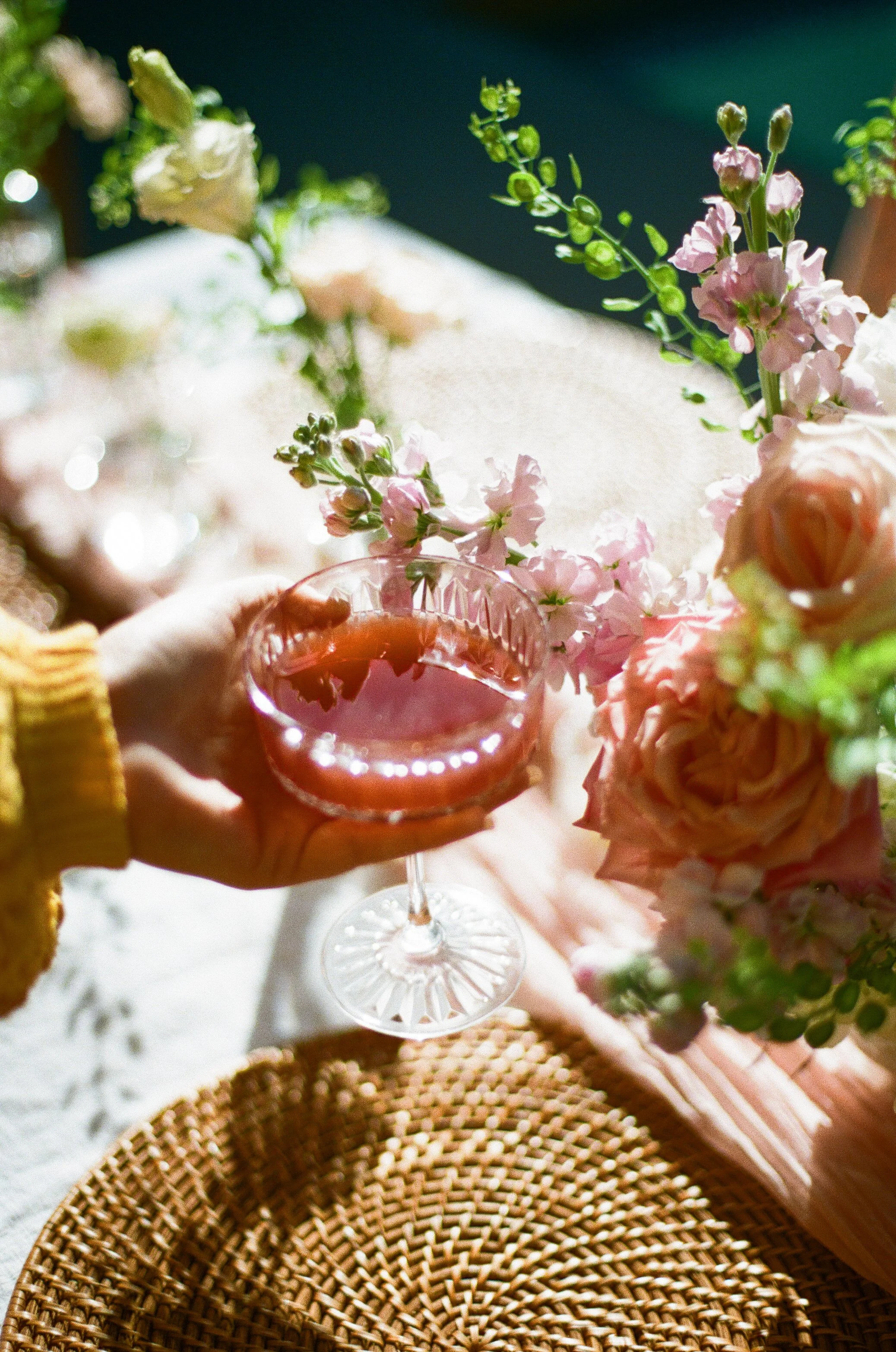 Person holding a glass of rosé wine surrounded by pink and white flowers on a table with woven placemat.