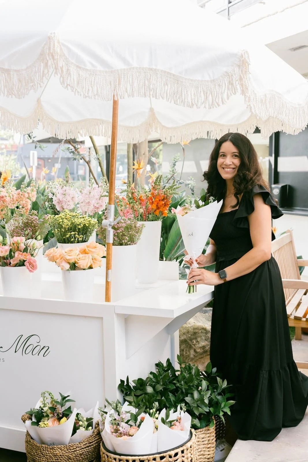 Woman in black dress with a bouquet, smiling at a flower stand with pink, yellow, and orange flowers under a white fringe umbrella.