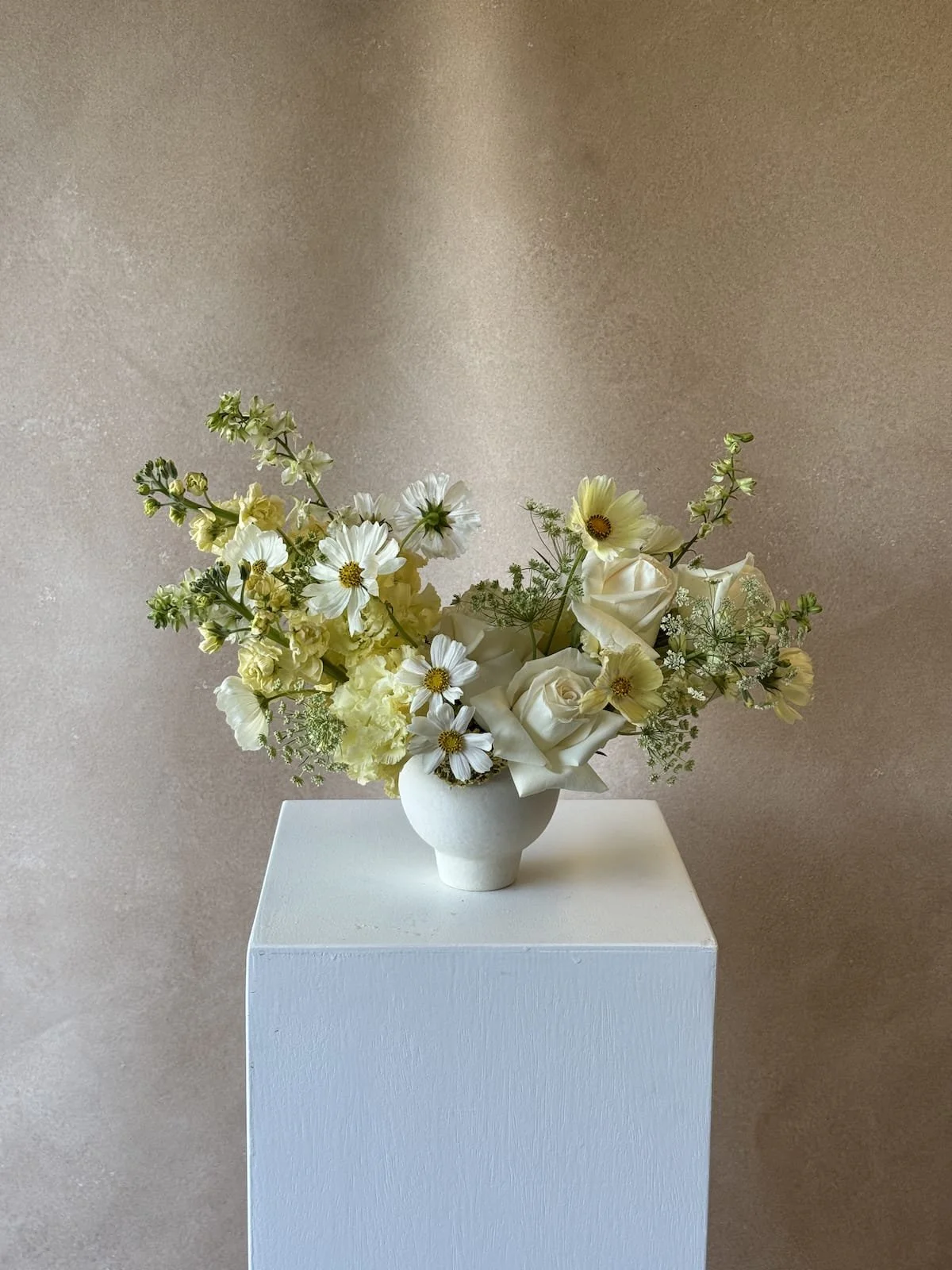 A white ceramic vase with a bouquet of white and light yellow flowers, including roses and daisies, placed on a white pedestal against a beige background.