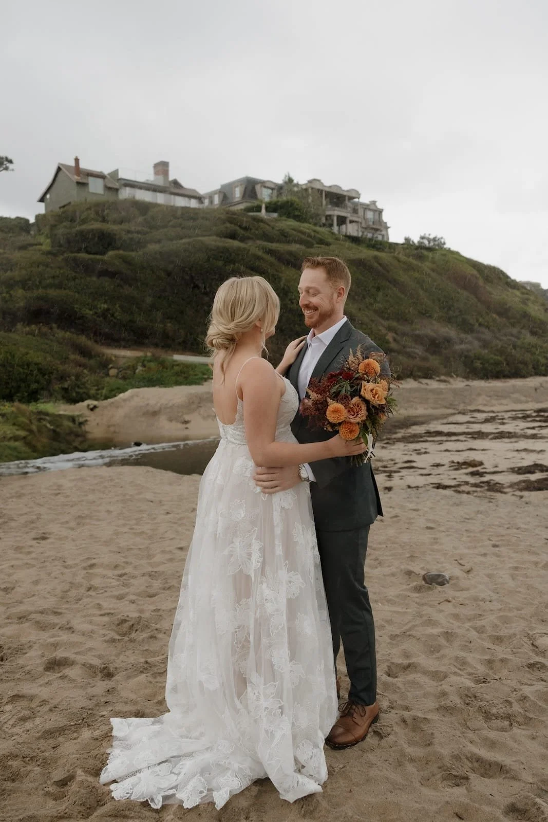 A bride and groom standing close together on a sandy beach, smiling at each other. The bride is holding a bouquet of orange and red flowers, wearing a white lace wedding dress. The groom is dressed in a dark suit with a white shirt, wearing brown shoes. In the background, there are green hills and large houses on the hilltop under a cloudy sky.