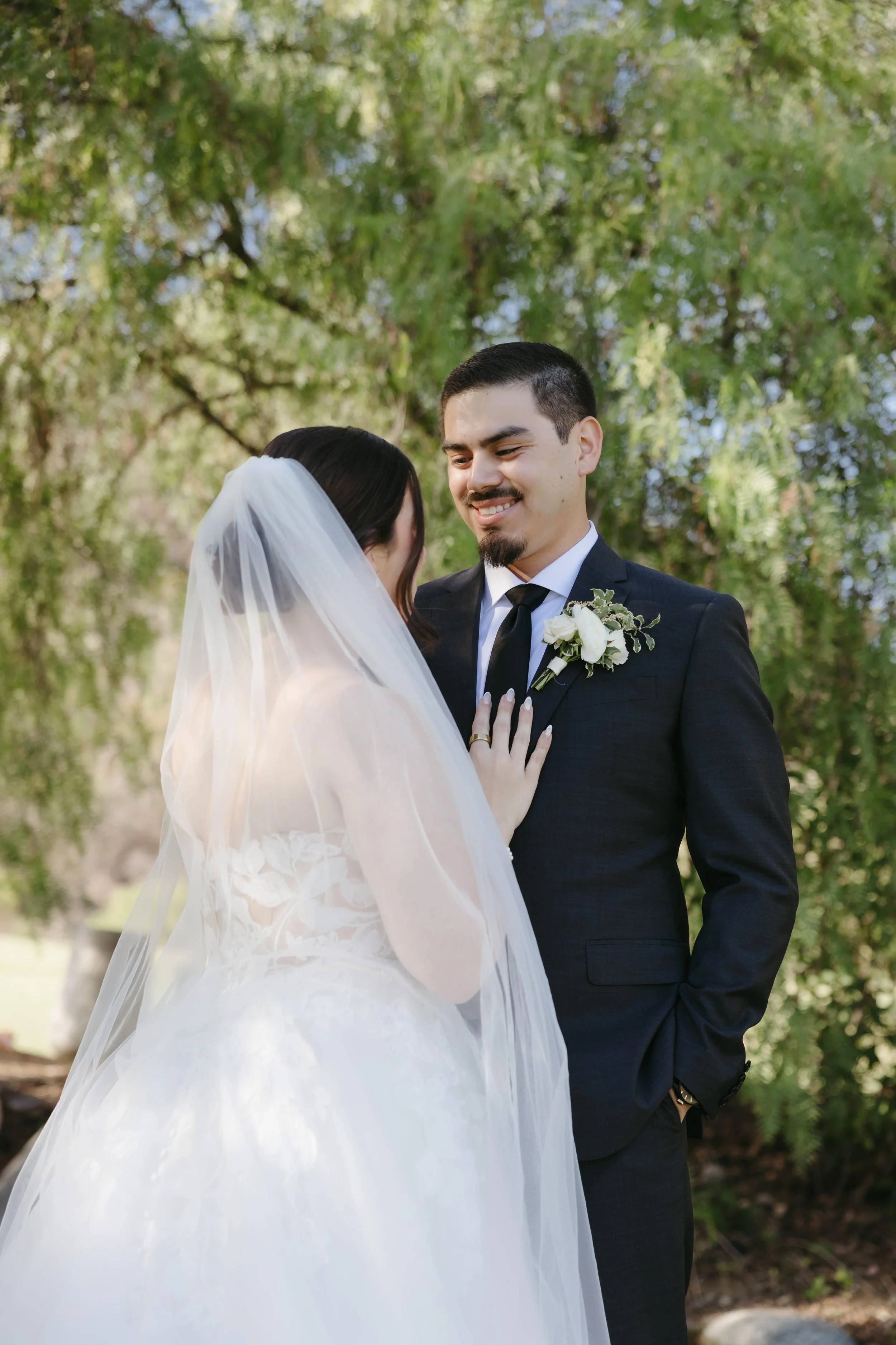 A bride and groom smiling at each other outdoors, with trees in the background. The bride wears a white wedding dress and veil, and the groom wears a dark suit with a boutonniere.