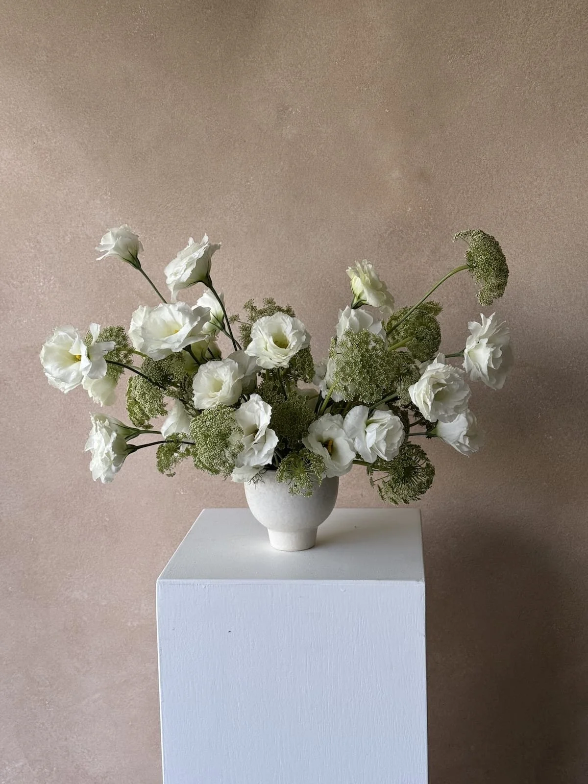 White floral arrangement in a white vase on a white pedestal against a beige wall.