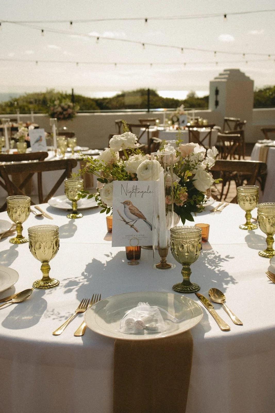 Elegant outdoor wedding reception table decorated with white and gold accents, floral centerpiece, gold glassware, and place settings, with string lights overhead and a scenic background of trees and water.