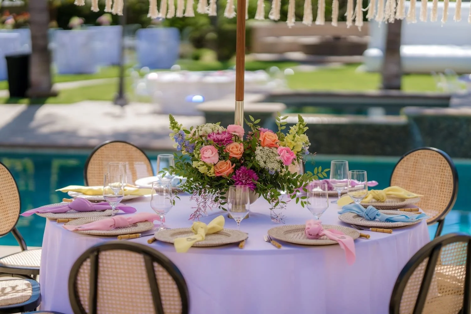 Round outdoor table set with pastel-colored napkins and glassware, decorated with a floral centerpiece under a large umbrella, near a pool.
