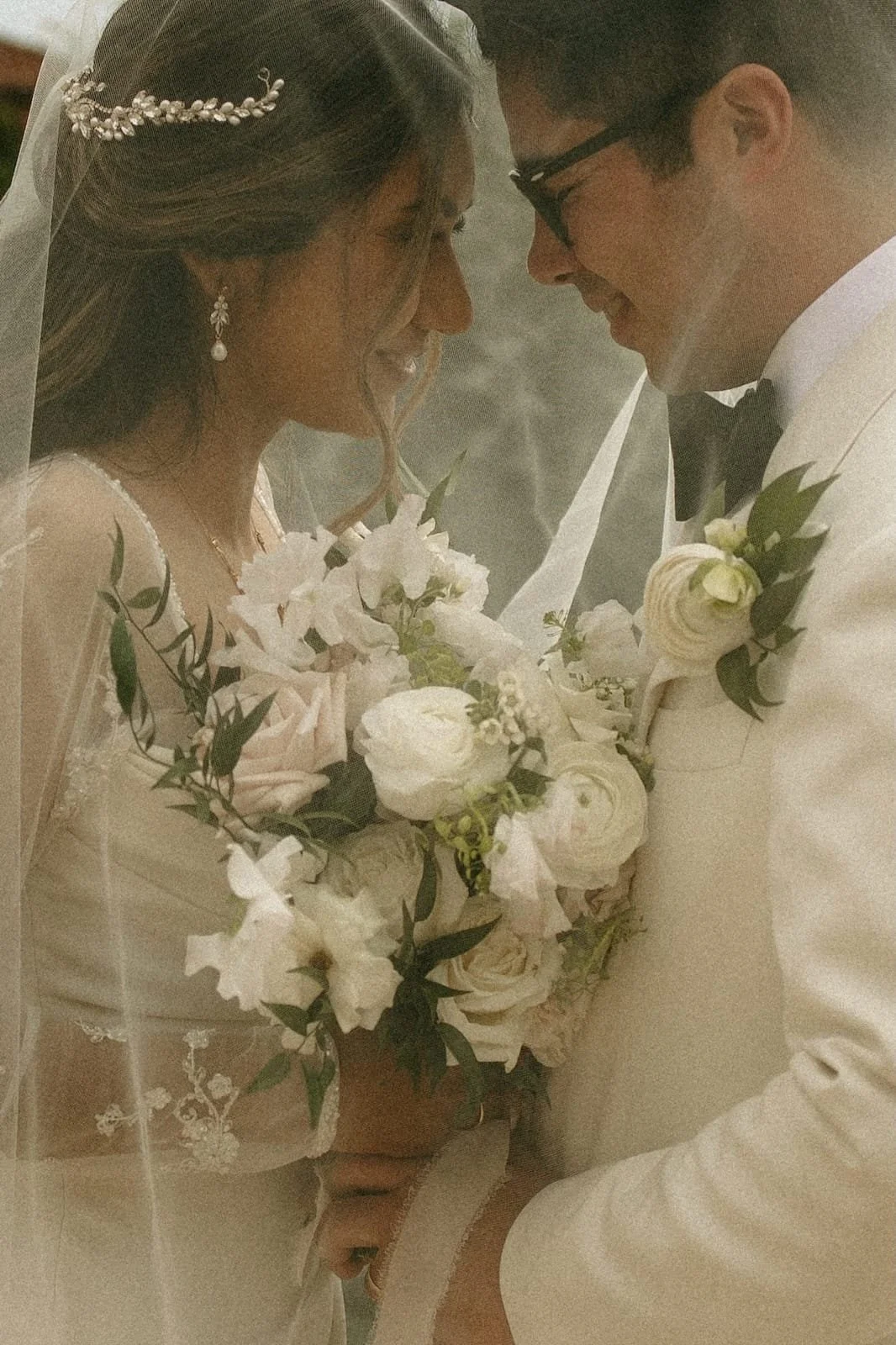 A bride and groom are close together, smiling and touching foreheads, with the bride holding a bouquet of white flowers and the groom dressed in a white tuxedo with a black bow tie.