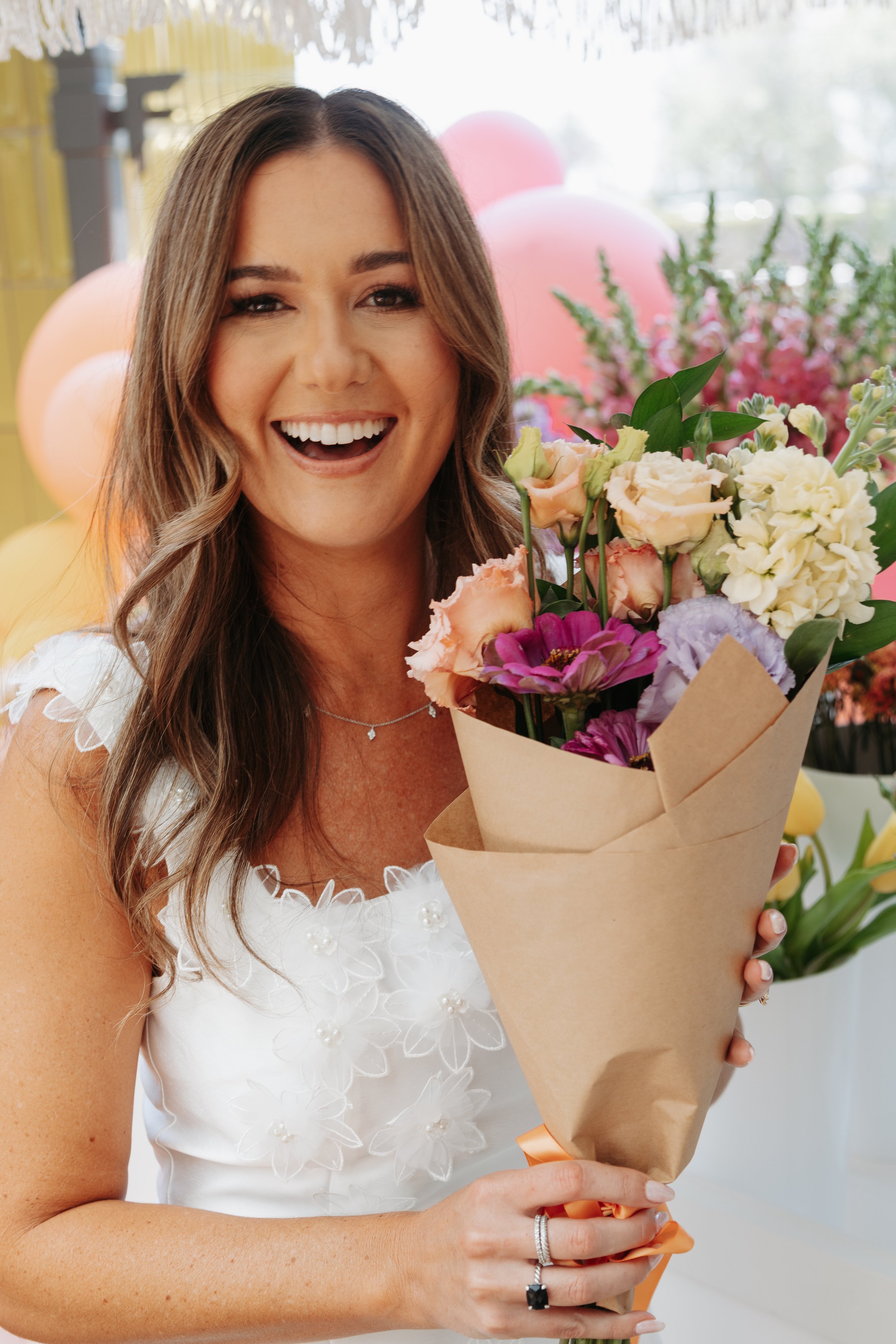 A woman with brown hair and a white dress holding a bouquet of colorful flowers, smiling at a celebration or event.