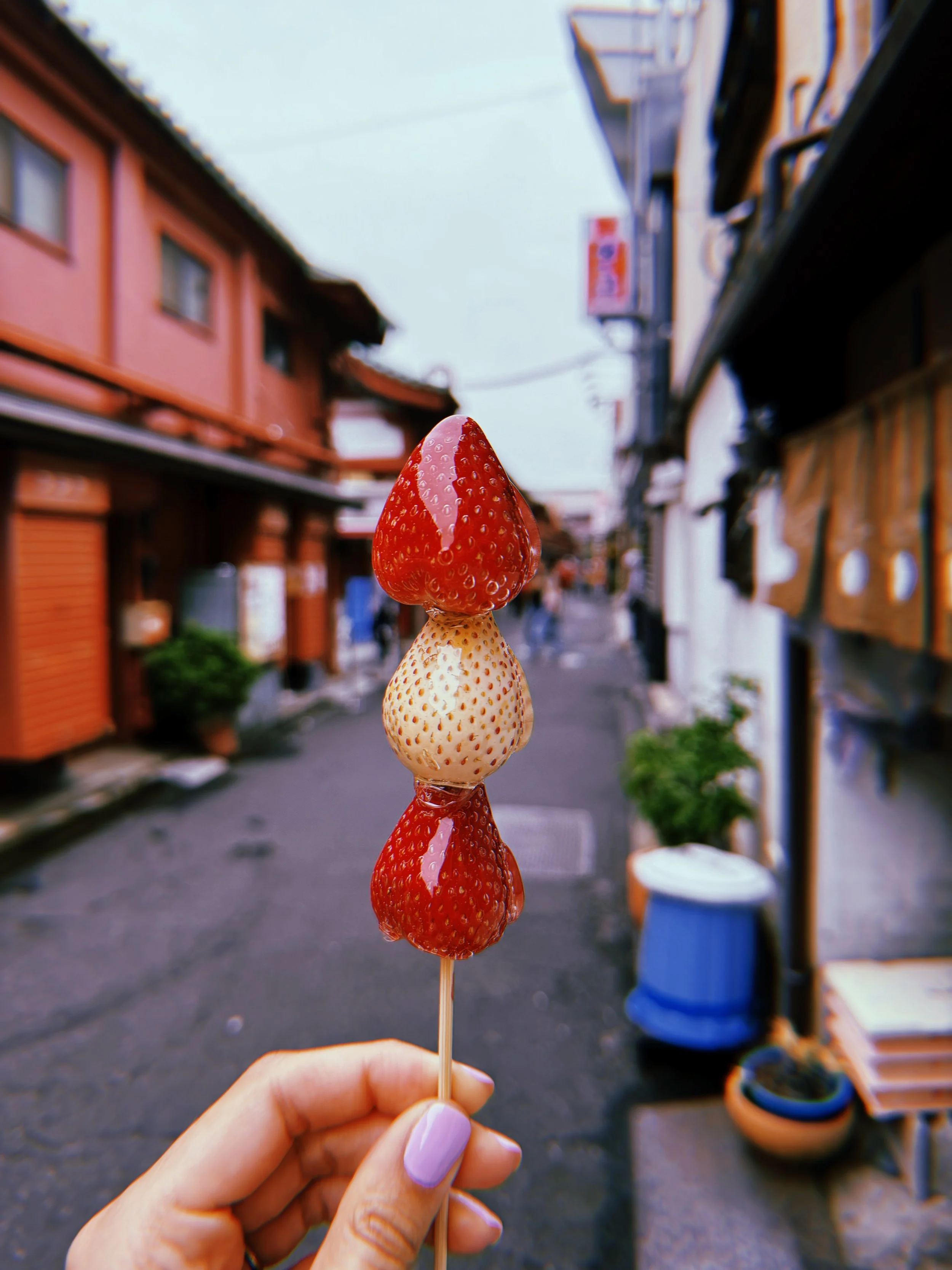 A hand holding a skewer with three strawberries, alternating between red and white, against a blurred outdoor street scene with traditional buildings.