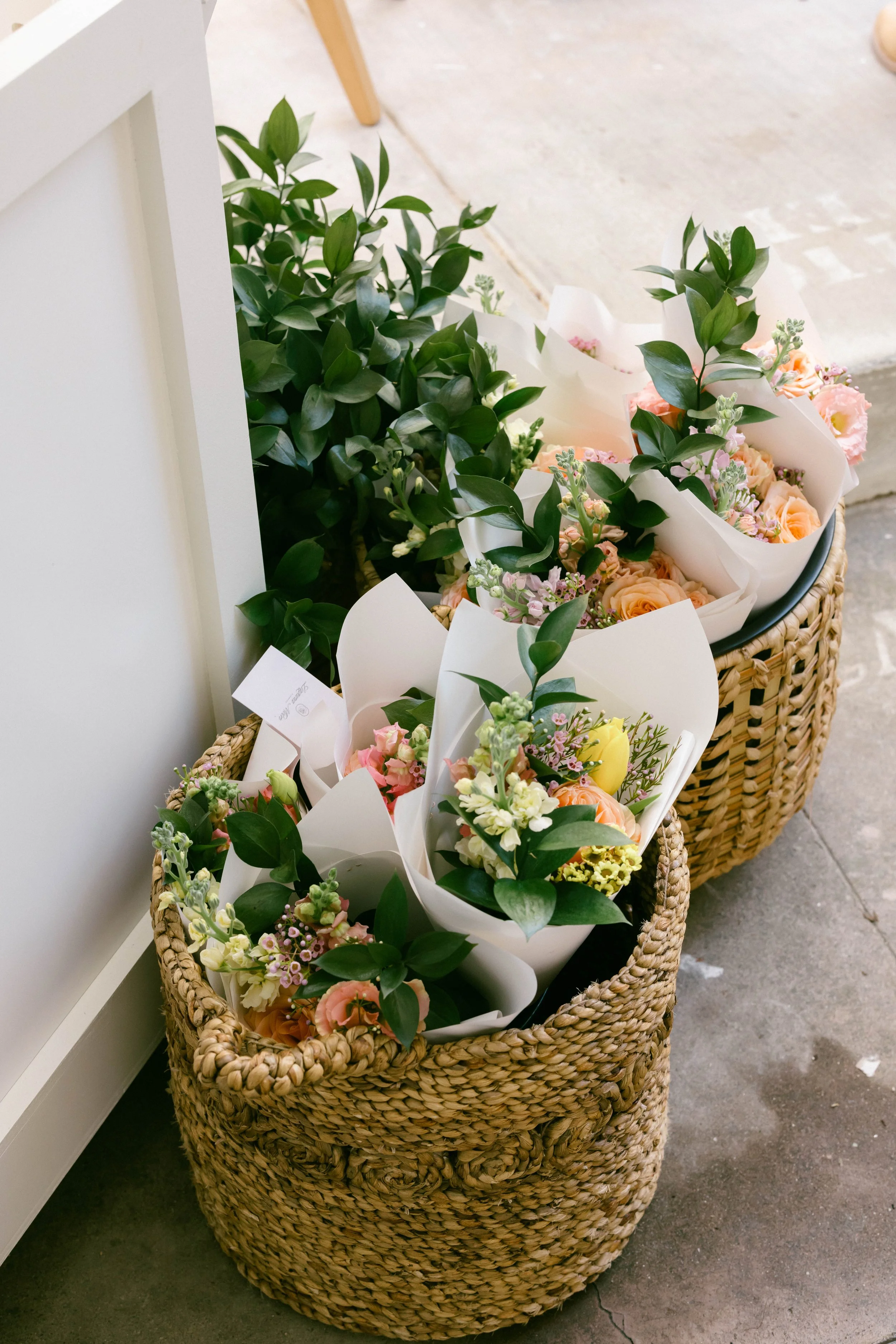 Wicker baskets filled with flowers and green foliage placed on the floor near a white wall.