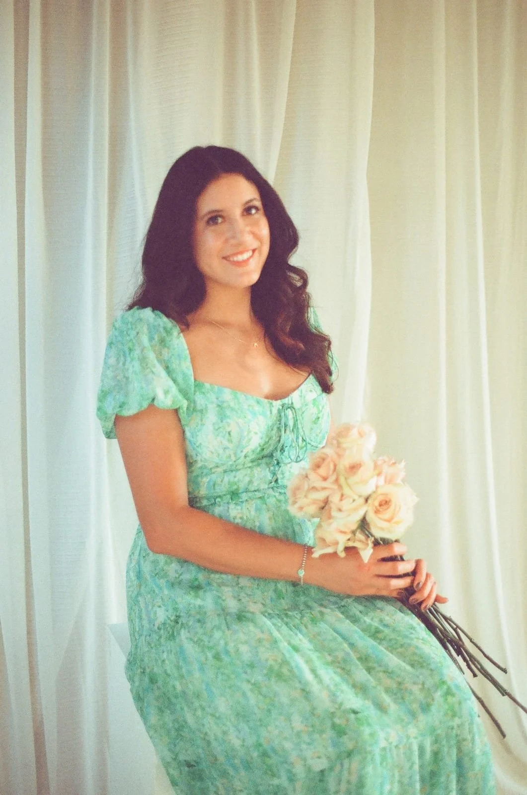 A woman with long dark hair wearing a light green floral dress, holding a bouquet of roses, sitting in front of cream-colored curtains, smiling at the camera.