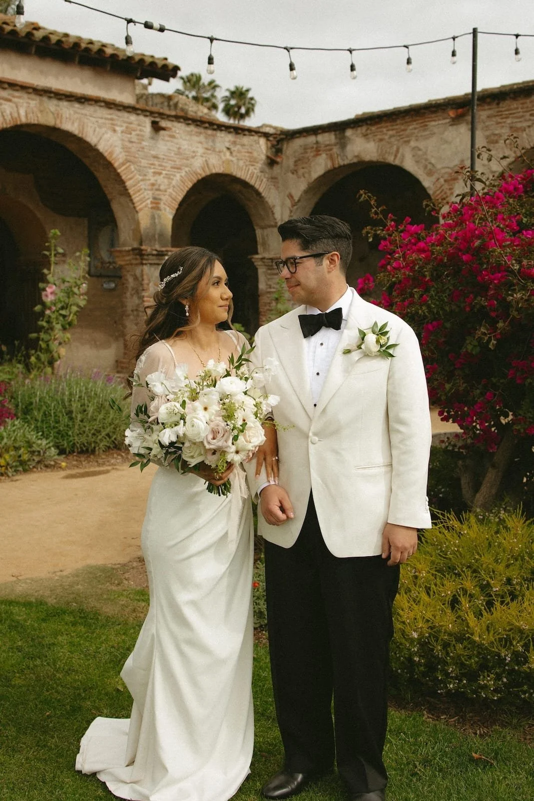 Bride and groom standing outdoors during their wedding, holding hands, with floral arrangements and a rustic building with arches in the background.
