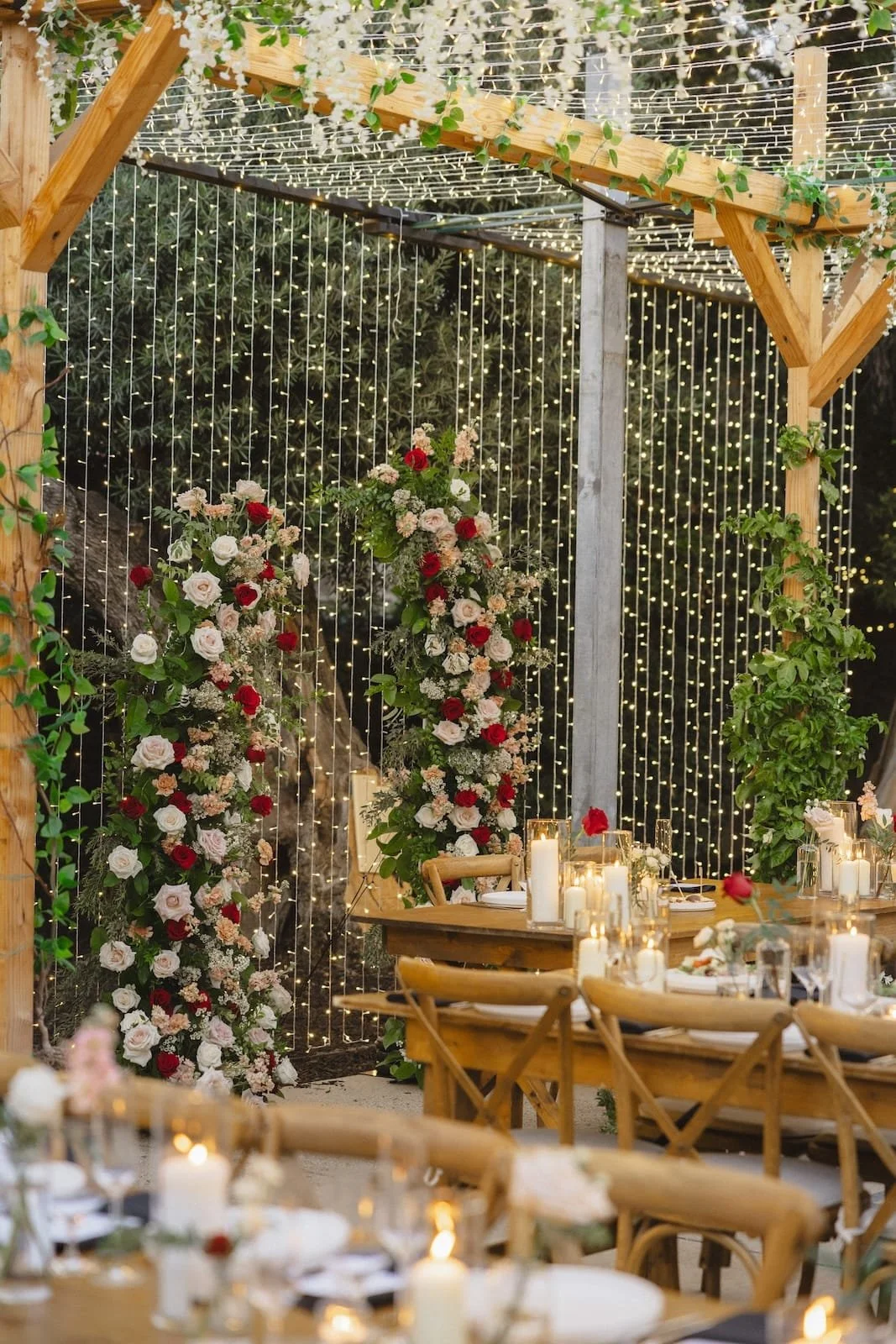 A decorated outdoor event space with string lights and floral arrangements on a wooden arch and tables, featuring candles and elegant table settings.