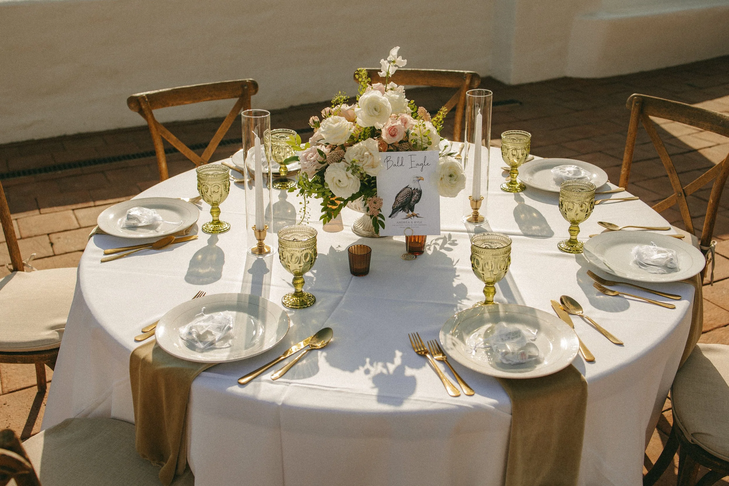 Elegant round dining table set for a formal event with a white tablecloth, gold utensils, and matching gold glasses. A floral centerpiece with white and pink flowers is accented with candles and a place card.