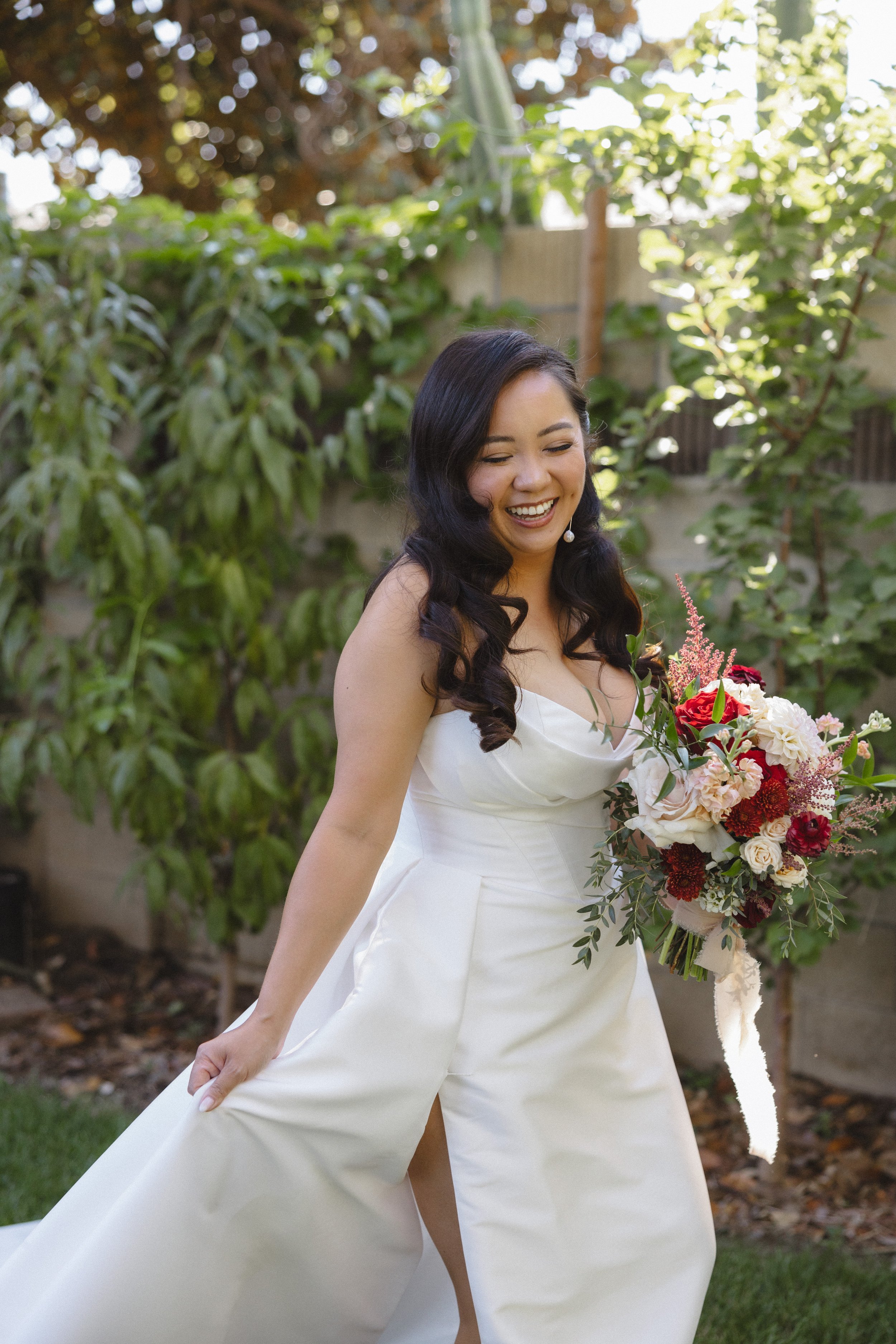 A smiling woman in a white wedding dress holding a bouquet in an outdoor garden setting.