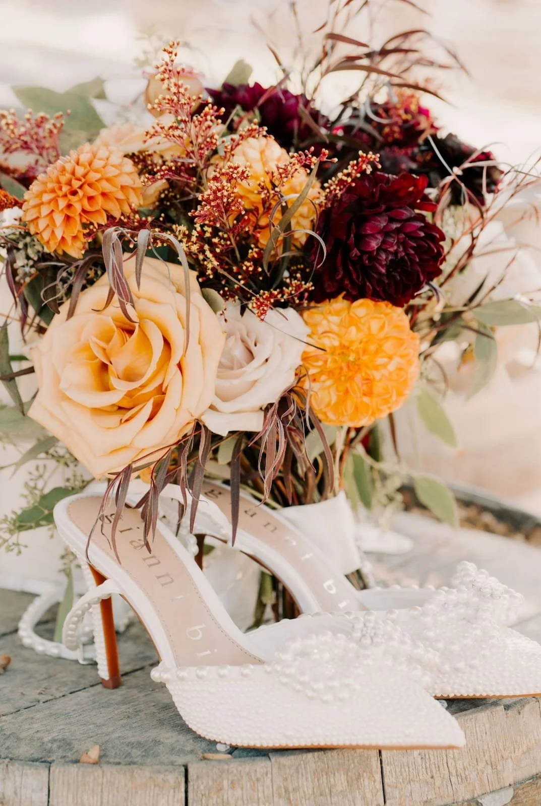 A bouquet of peach, white, orange, and burgundy flowers with greenery, placed behind a pair of white high-heeled shoes decorated with beads, on a wooden surface.