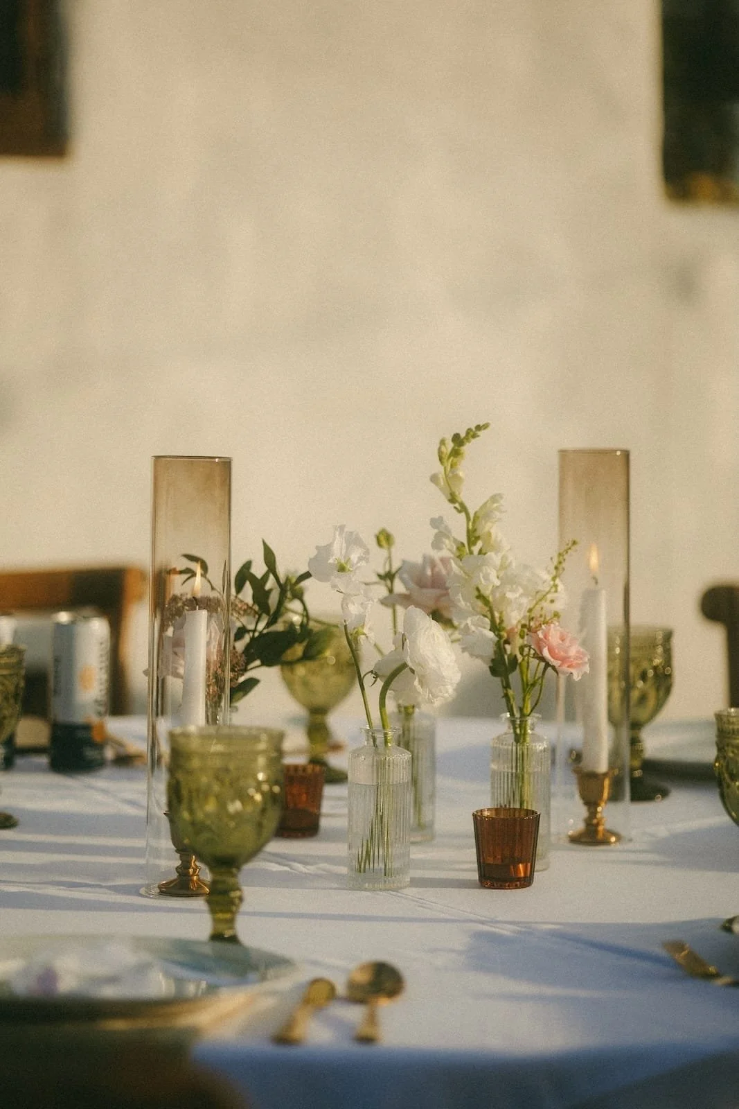 Table decorated with glass vases holding white and pink flowers, tall candle holders with candles, and colored glassware, with sunlight casting shadows.