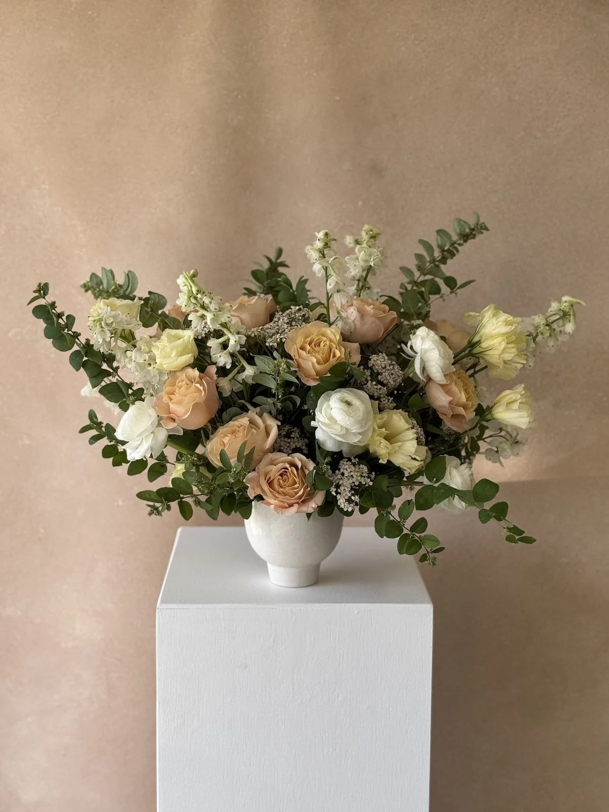 A floral arrangement with cream, peach, and white flowers in a white vase on a white pedestal against a beige background.