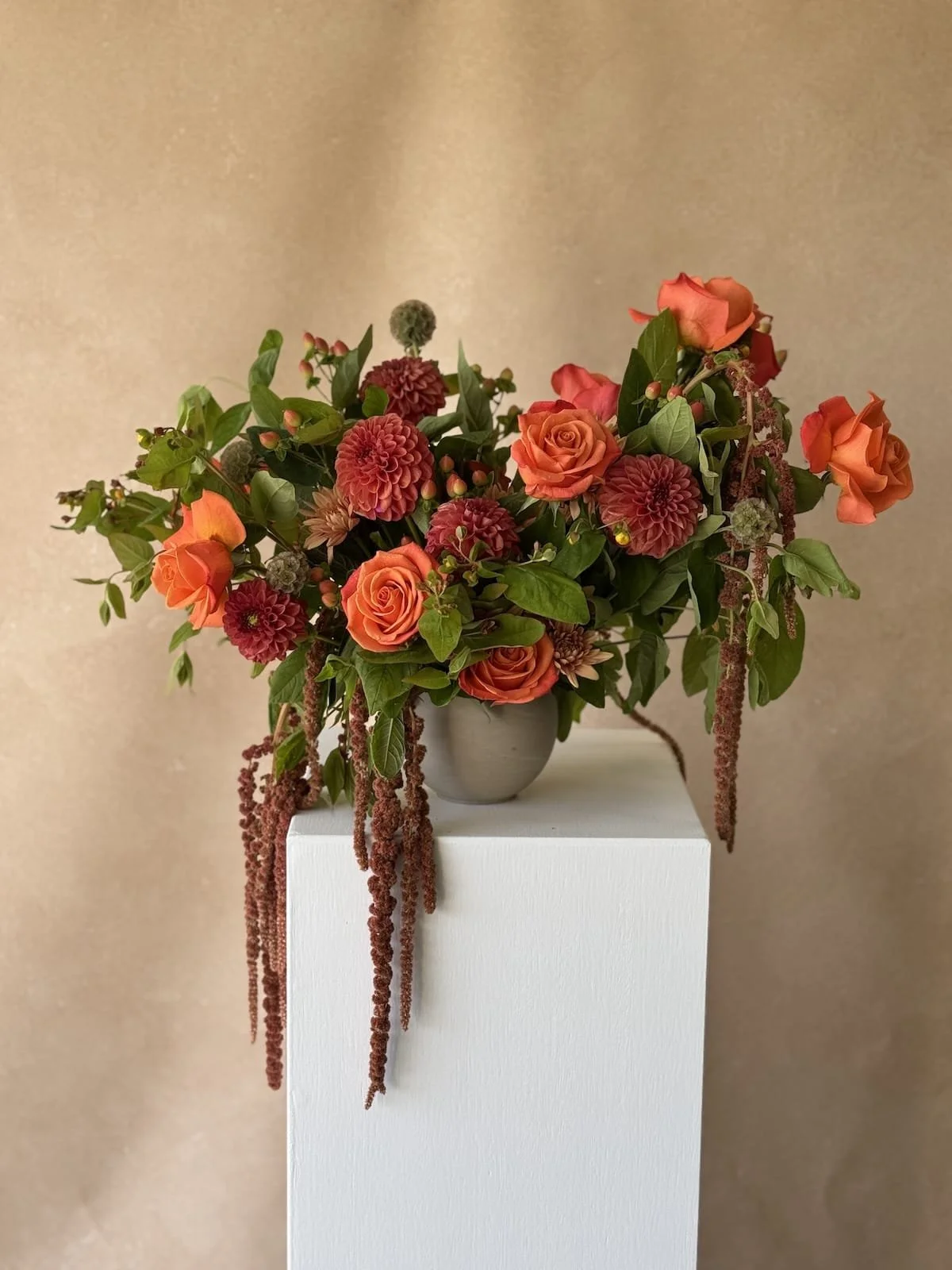 A floral arrangement with orange roses, burgundy dahlias, and hanging amaranthus in a grey vase on a white pedestal against a beige background.