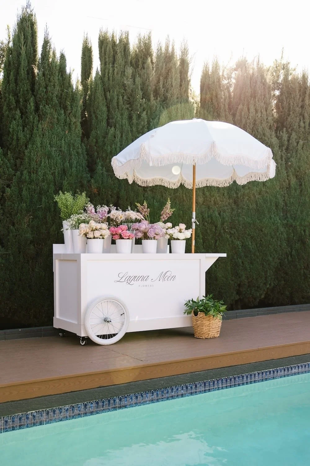 A white flower cart with flowers and a white parasol, with 'Laguna Moon Flowers' written on it, next to a swimming pool and green trees in the background.