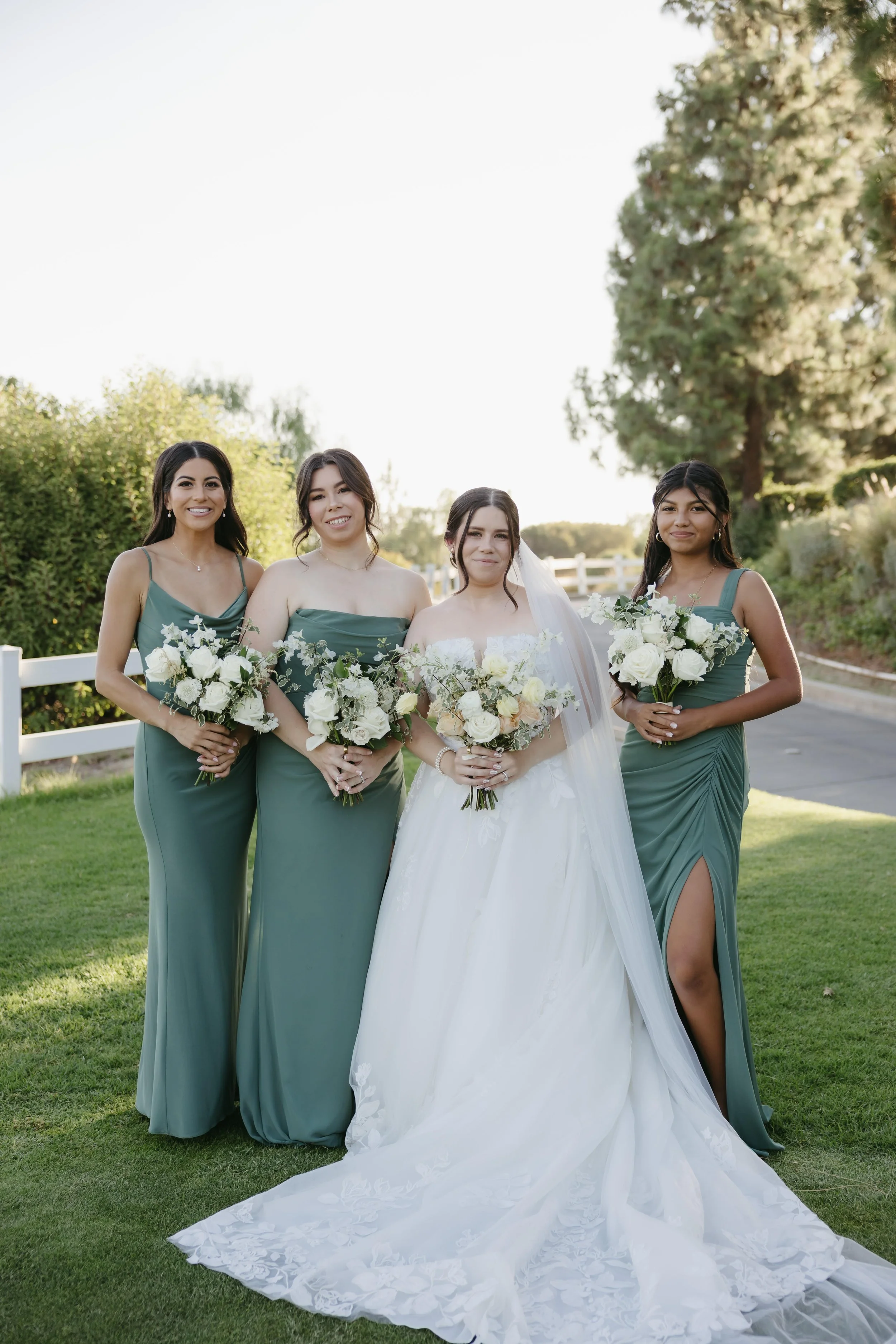 A bride in a white wedding gown with a long train poses outdoors with three bridesmaids in matching green dresses, all holding bouquets of white roses and greenery, on a grassy area with trees and a white fence in the background.
