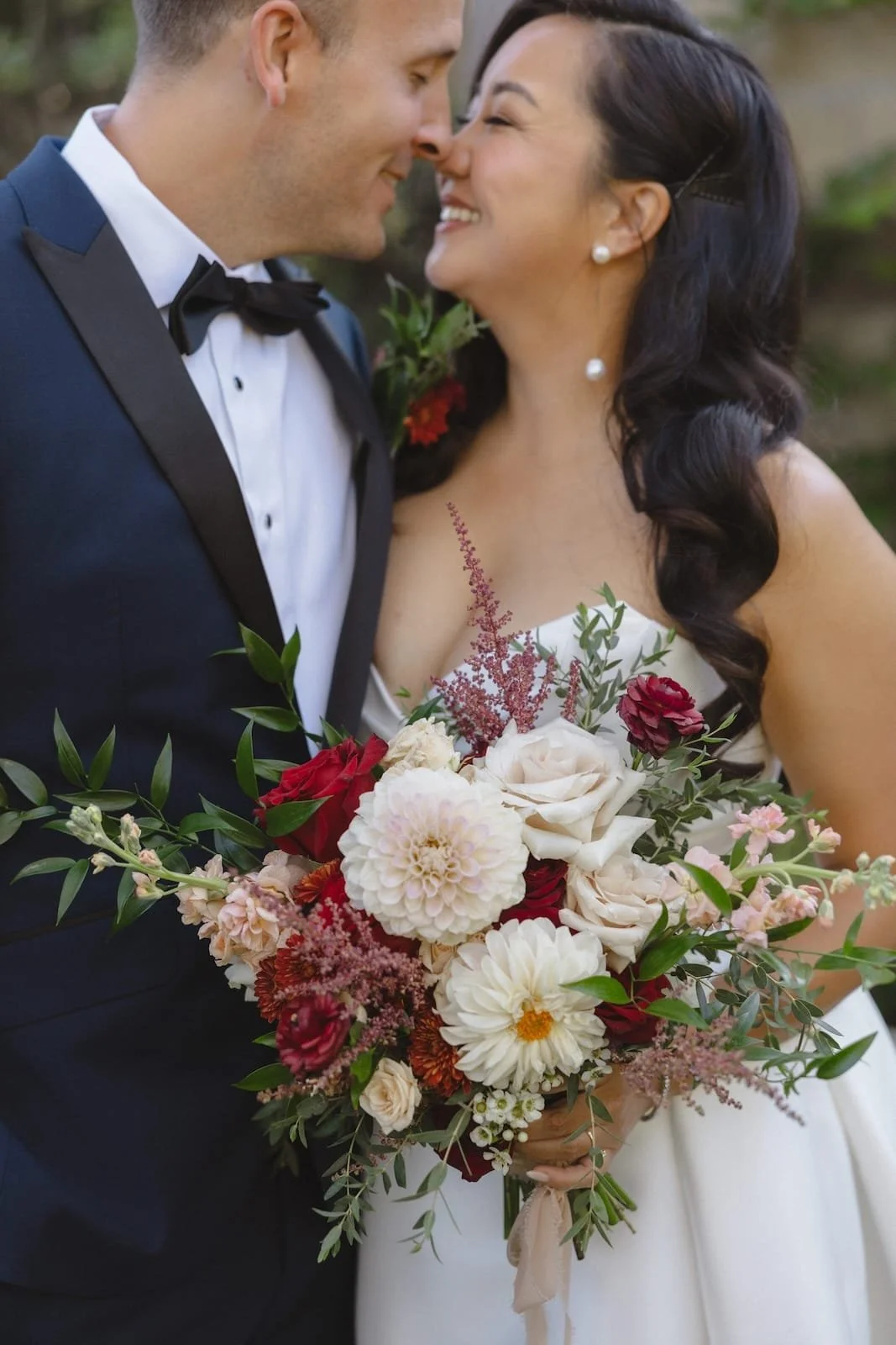 A newlywed couple is close together, smiling and nearly touching noses. The bride is holding a large, colorful bouquet of flowers, including white, red, and pink blossoms with green foliage. The groom is wearing a dark tuxedo with a bow tie, and the 