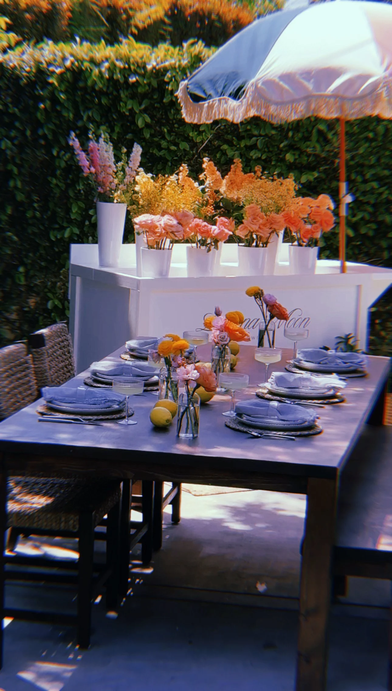 An outdoor dining table set for a meal, decorated with flowers in vases, lemons, and glasses, surrounded by wicker chairs. Behind the table, a white cart displays additional flower arrangements under a large umbrella with a fringed edge.