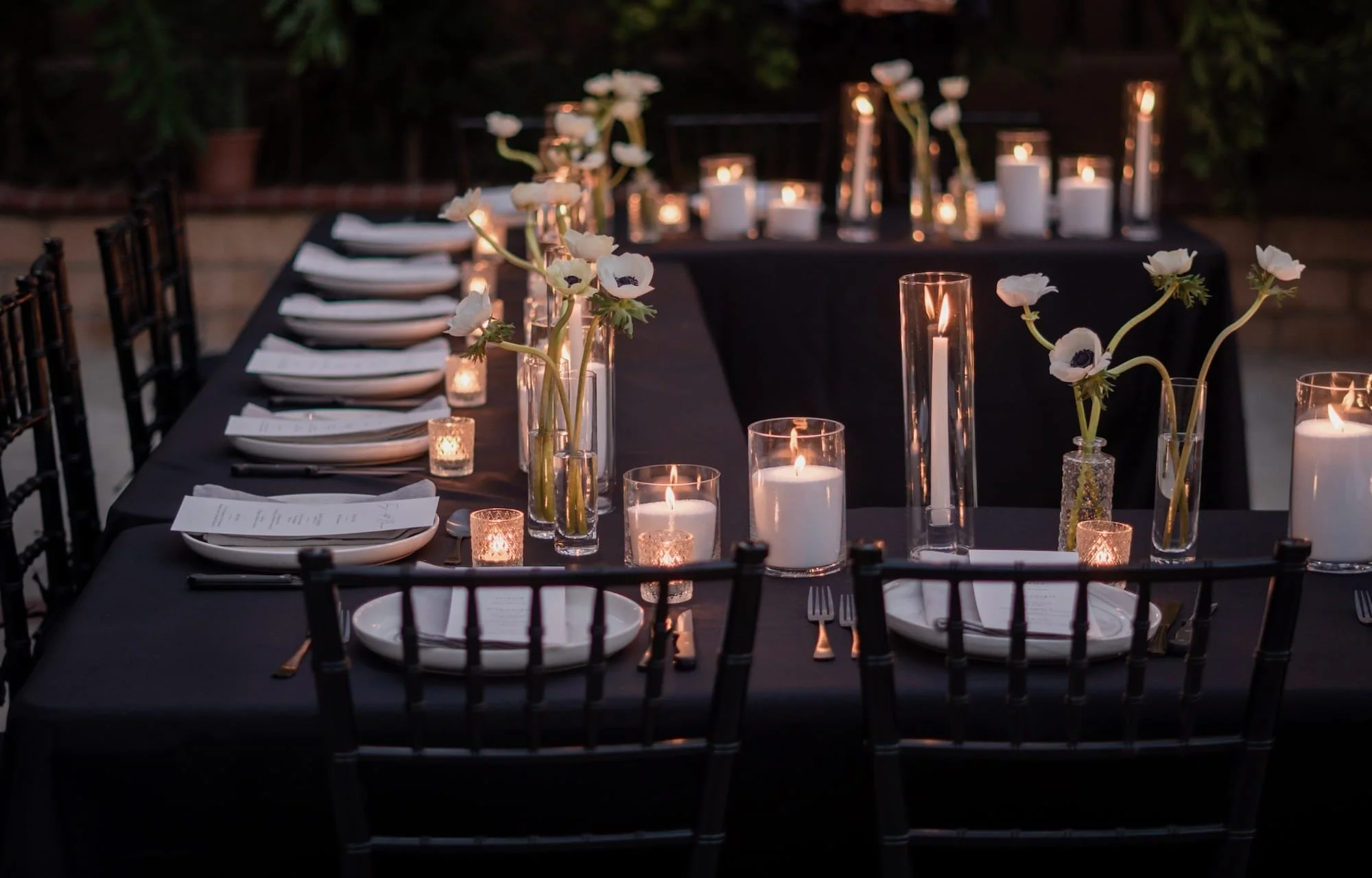 An elegant outdoor dinner table decorated with white flowers in glass vases, lit candles, and black tablecloths, set for a formal event at dusk.